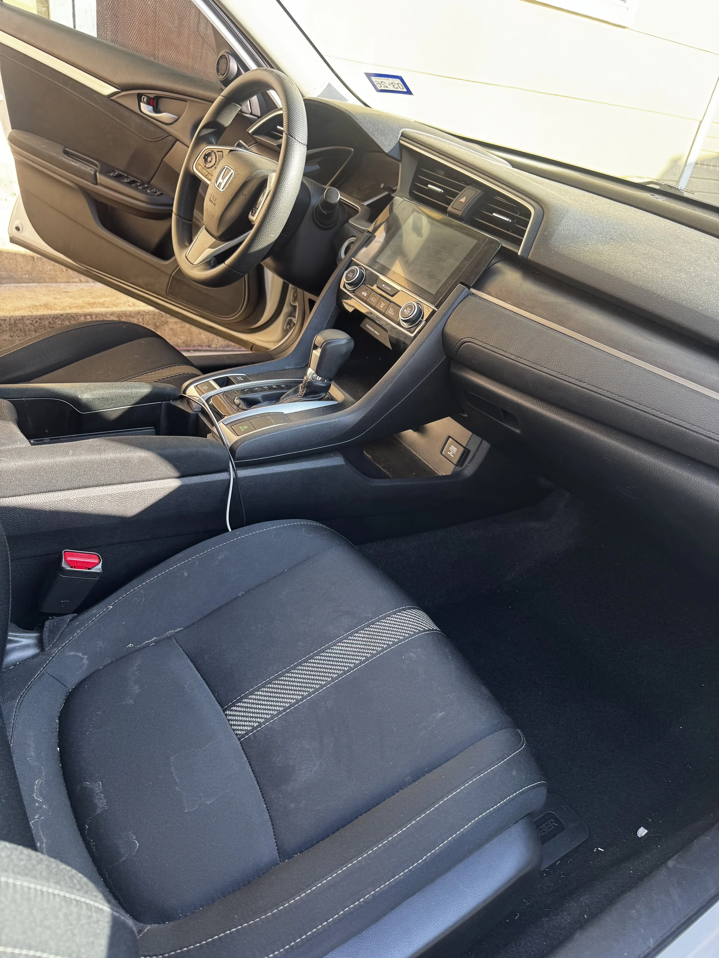 Inside a car, focusing on the front passenger seat with black fabric upholstery featuring light stitching, and the center console with controls and gear shift.