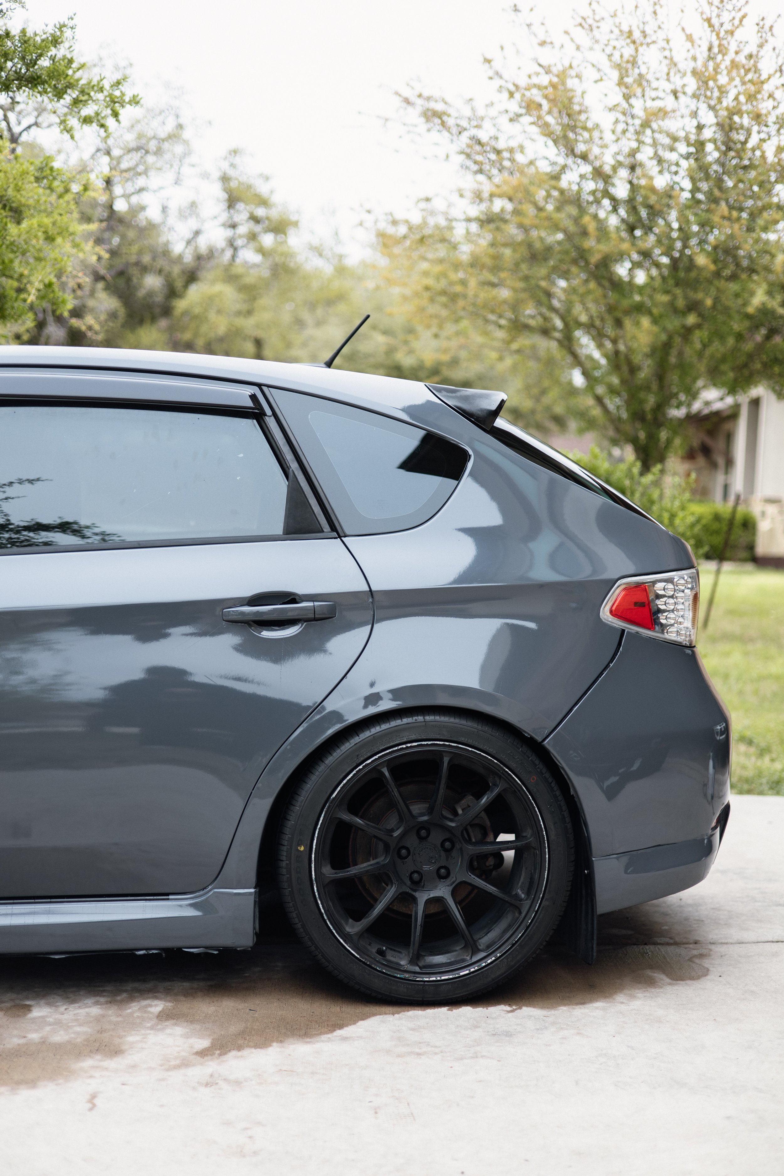 Close-up of a gray sports car parked outdoors with black rims, part of a tree and house visible in the background.