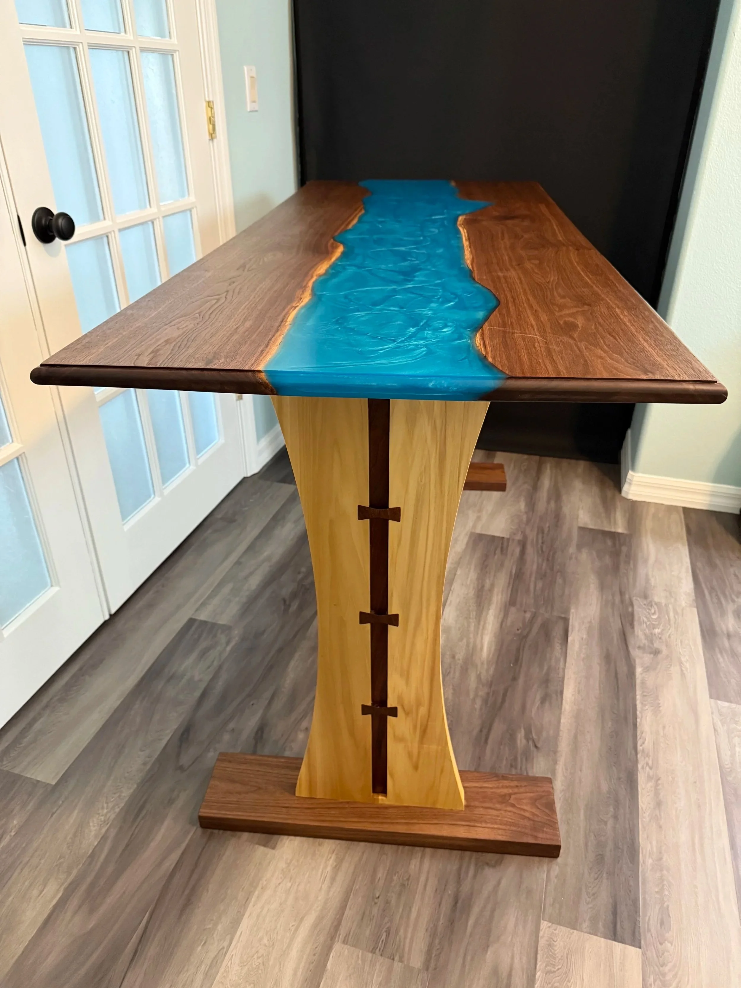 A black walnut wooden table with a river-shaped epoxy resin inlay in the center, supported by a wooden base with a dark finish.