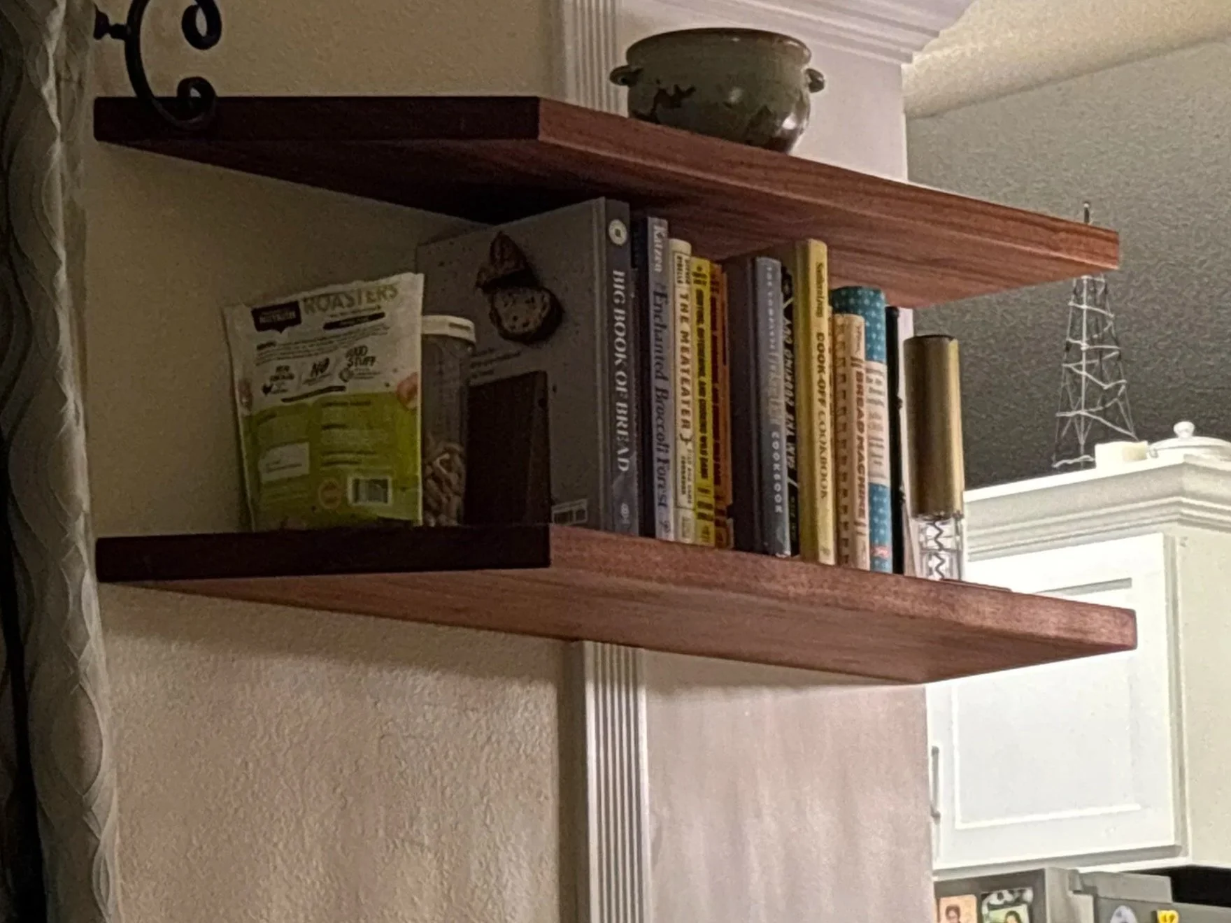 A wooden bookshelf with two shelves filled with books, a jar of snacks, and a decorative clock. Located near a window with a white frame in a room with beige walls.