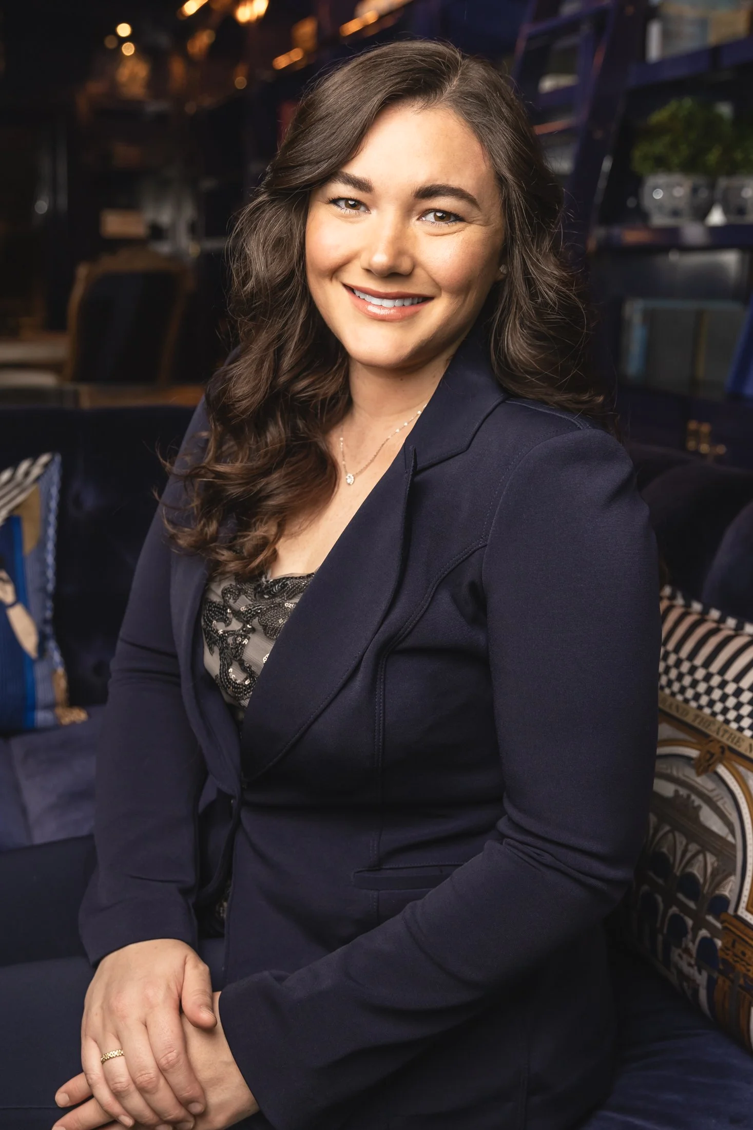 A woman with long, wavy brown hair, wearing a navy blazer and a black lace top, smiling while seated indoors with a dark background and patterned cushions.
