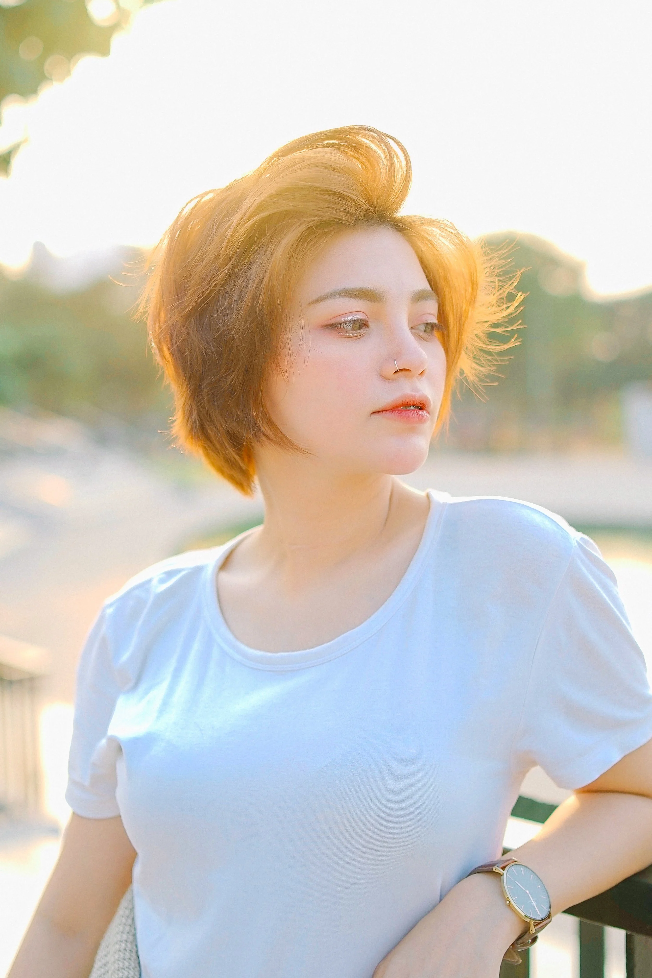 A young woman with short, tousled red hair wearing a white T-shirt and a wristwatch, standing outdoors in sunlight with a blurred background of trees and sky.