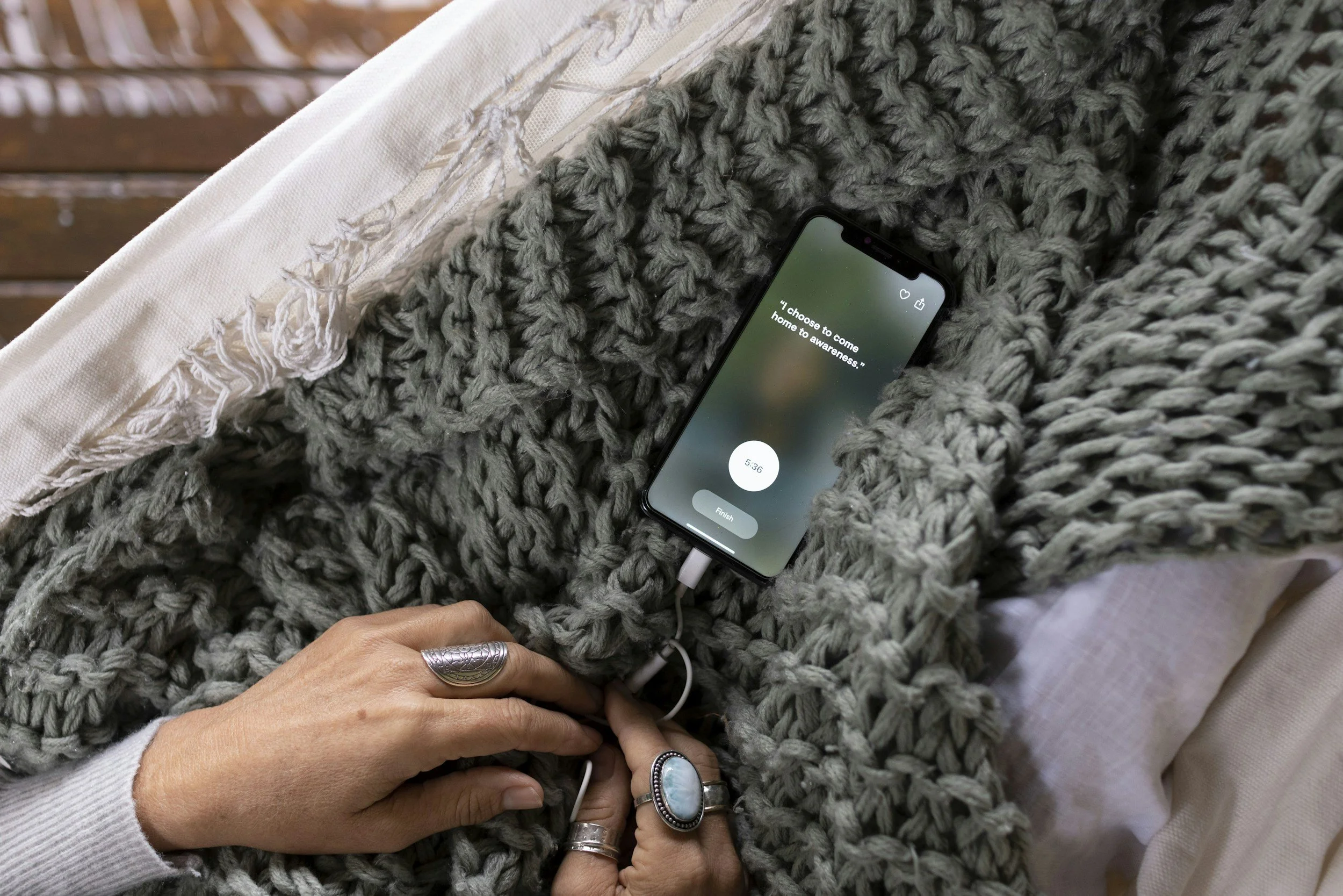 A person with rings on their fingers is lying in bed, covered with a large chunky gray knit blanket. They are holding a phone connected to a charger, displaying an app with a quote about choosing to come home to awareness.
