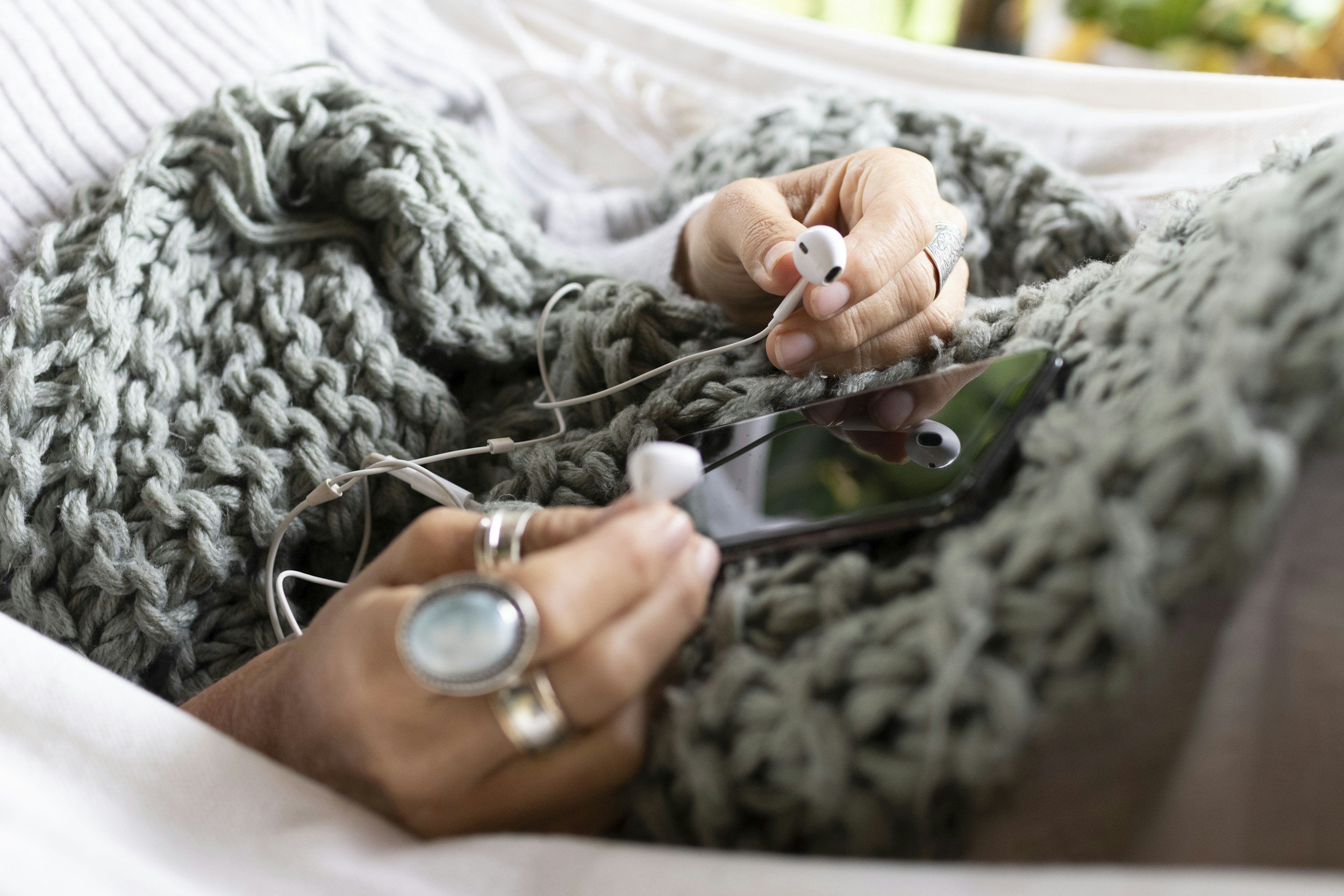 A person holding a smartphone with earphones plugged in, resting on a chunky knit blanket.