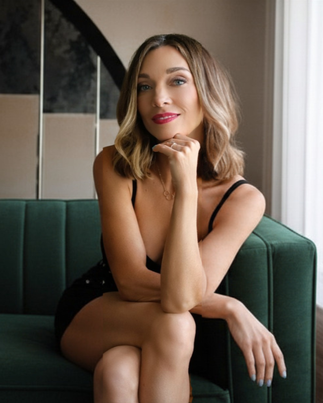 Elisha Lee with shoulder-length light brown hair, wearing red lipstick and a black dress, sitting on a green sofa near a window.