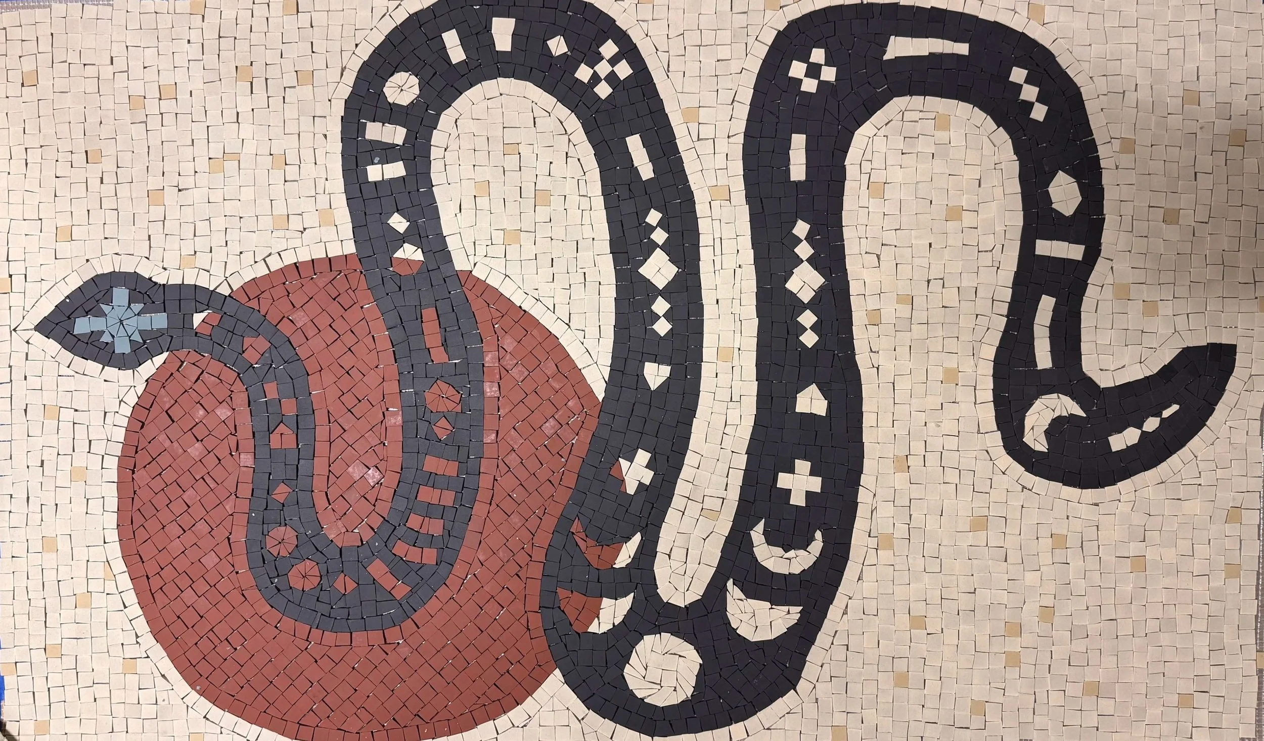 Colorful mosaic artwork featuring a red sun with black snake designs and various patterns, in progress on a worktable surrounded by mosaic tiles and tools.