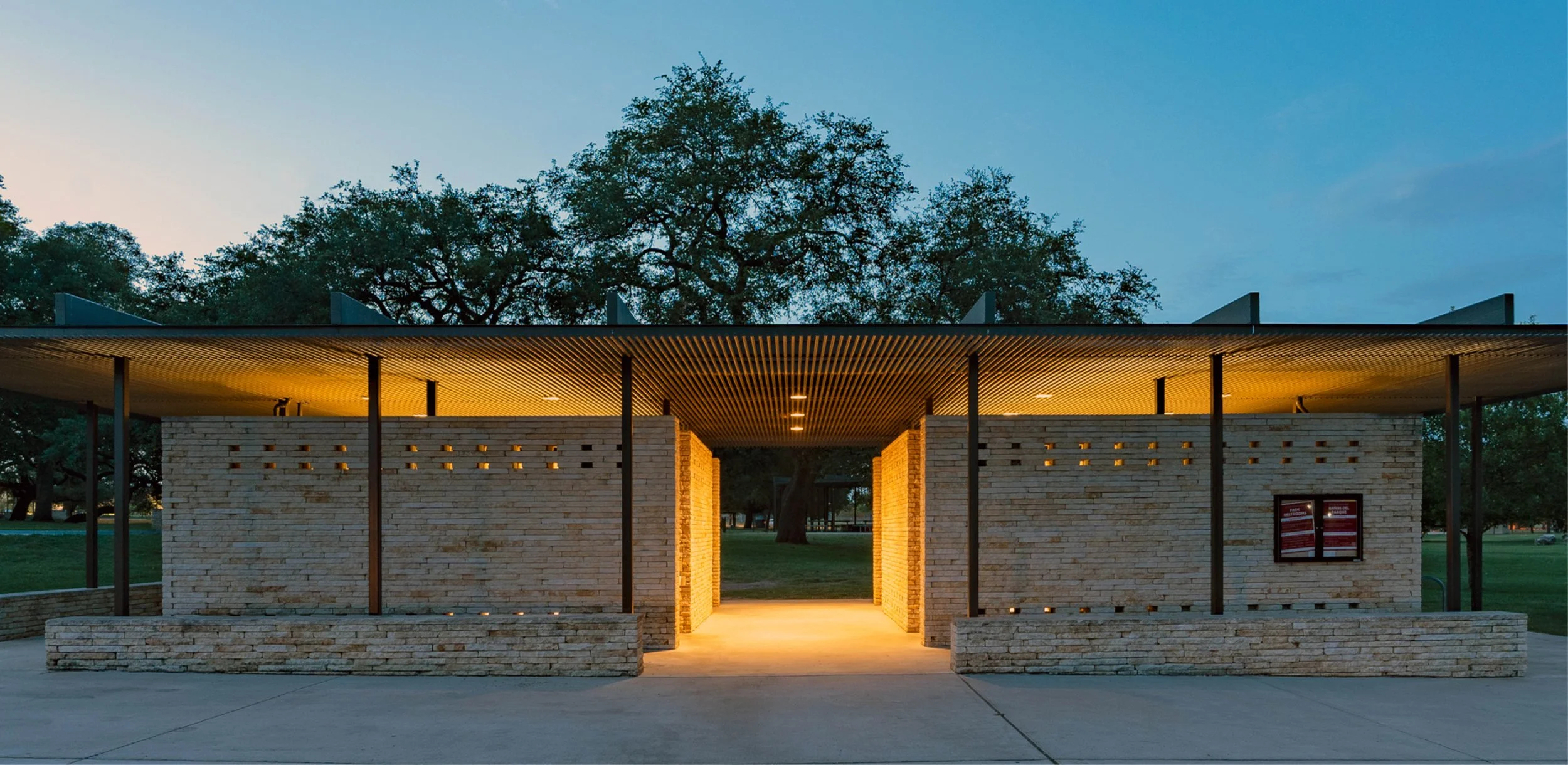 Modern park restroom at San Gabriel Park in Georgetown Texas, with brick walls and a slatted wooden roof, lit by warm lighting, set in a park with trees in the background at dusk.