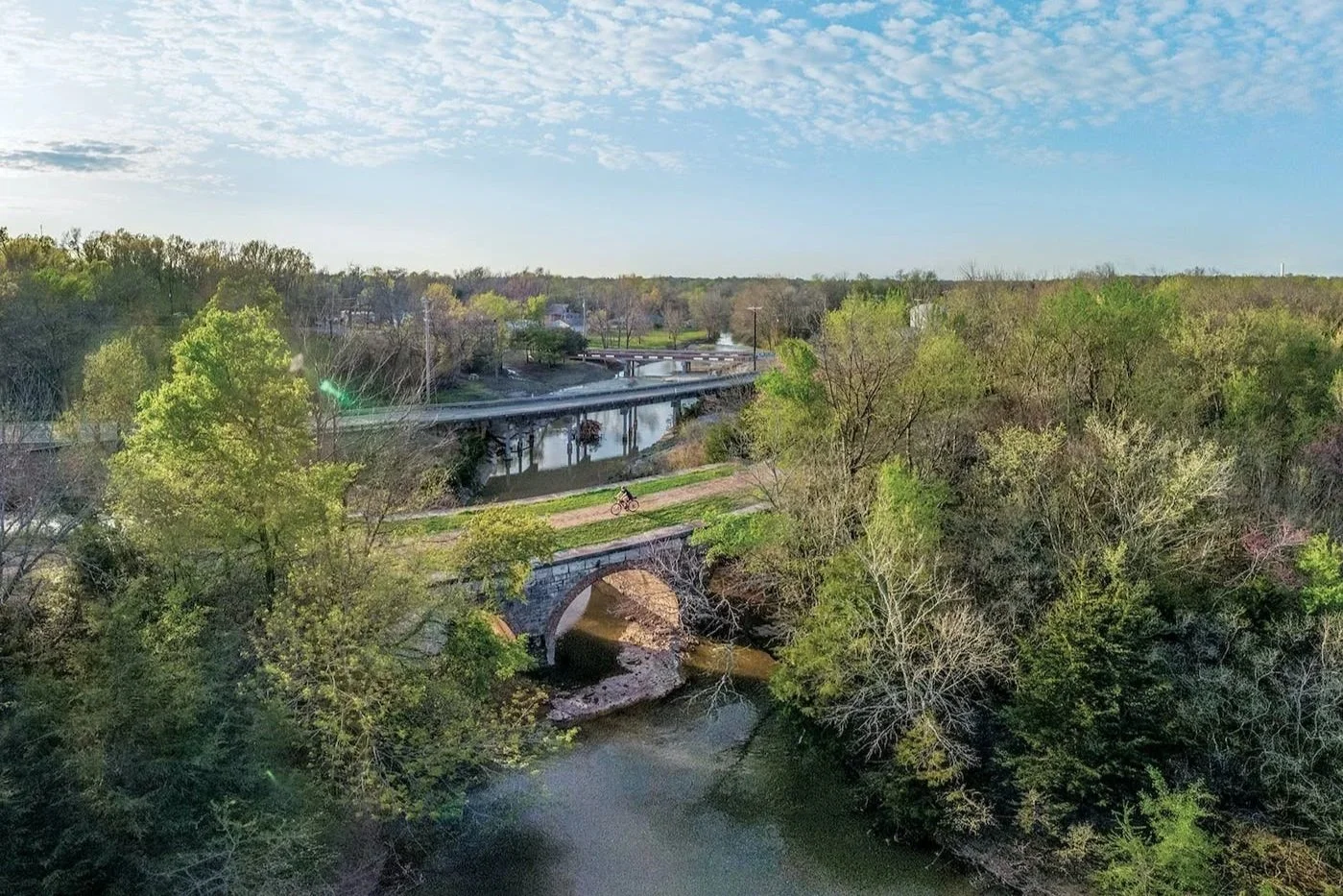 Aerial view of a river with green trees, a stone bridge, and a person riding a bicycle along a dirt path. There are bridges and a partly cloudy sky in the background.
