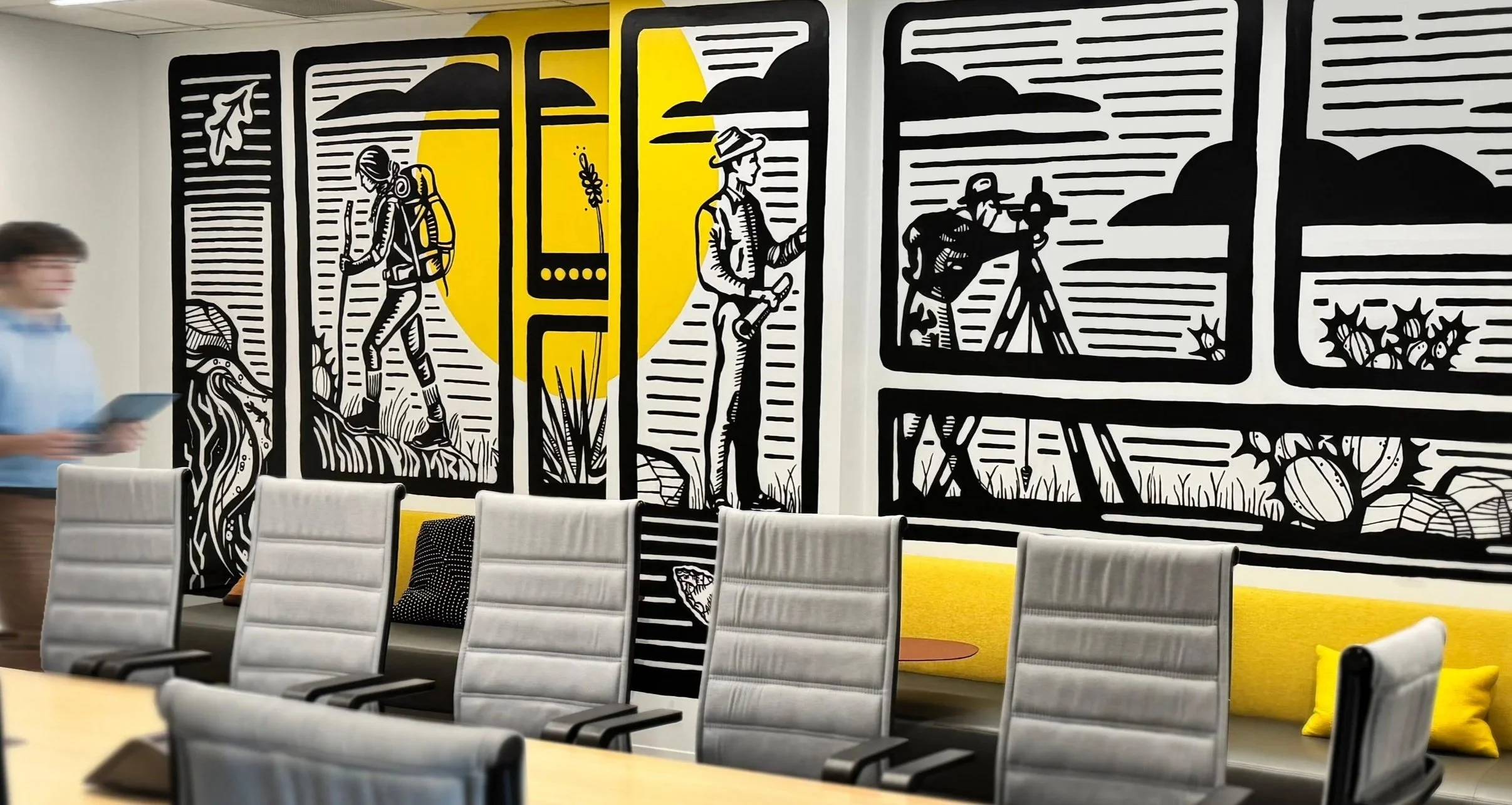 Modern conference room with a wall mural featuring black and white line art of outdoor scenes and a yellow background. There are gray conference chairs, a yellow padded bench with pillows, and a blurred person holding a tablet.