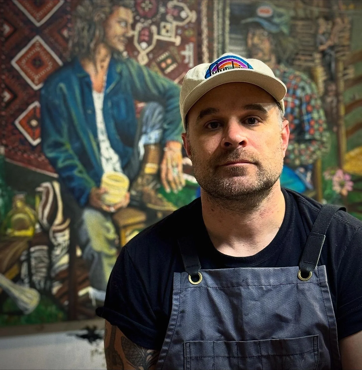 Photograph of Drew Carman wearing a white cap with a rainbow logo, a black t-shirt, and an apron in front of a colorful mural of a woman with long hair sitting among traditional and cultural items.