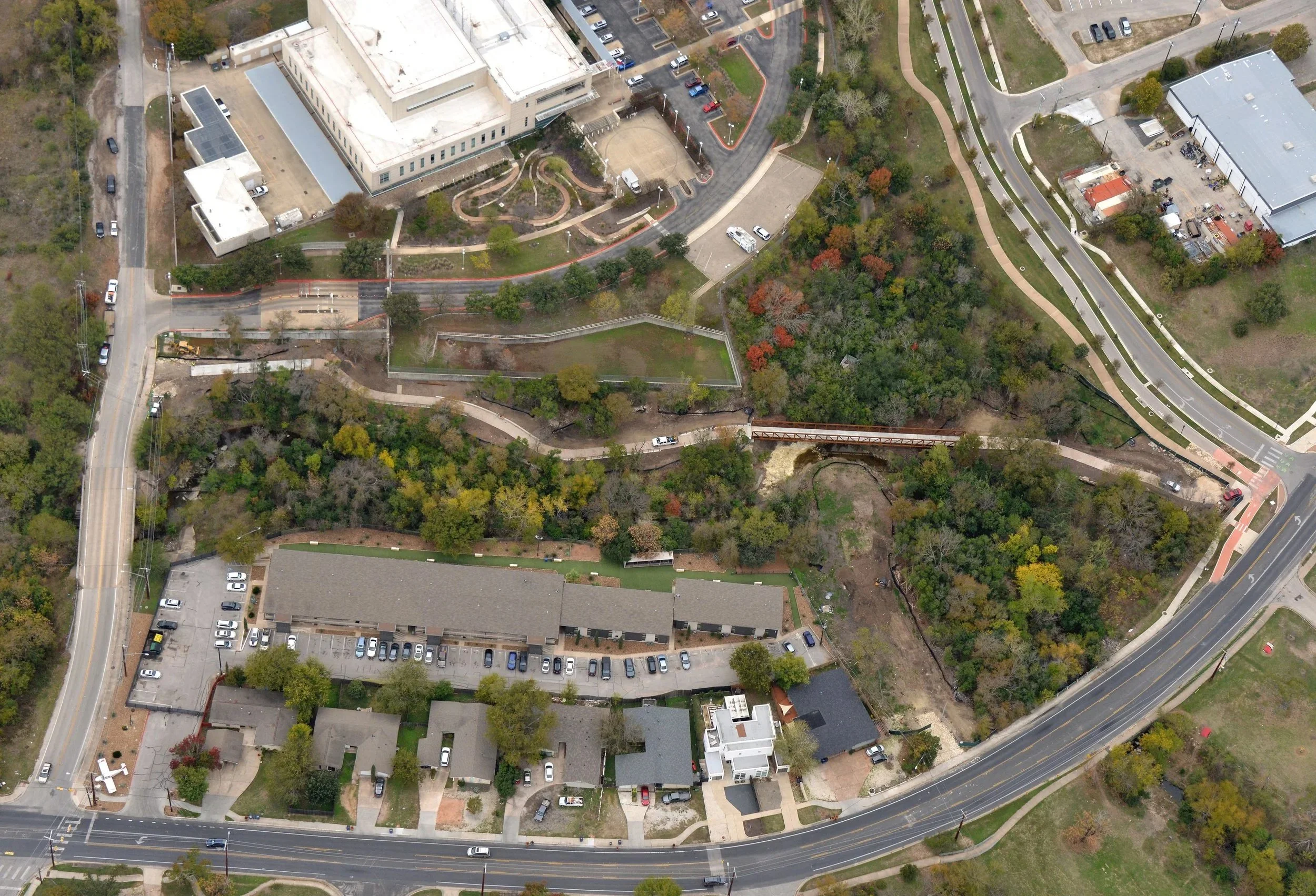 An aerial view of an urban area with buildings, parking lots, roads, and green spaces, some with walkways and trees.