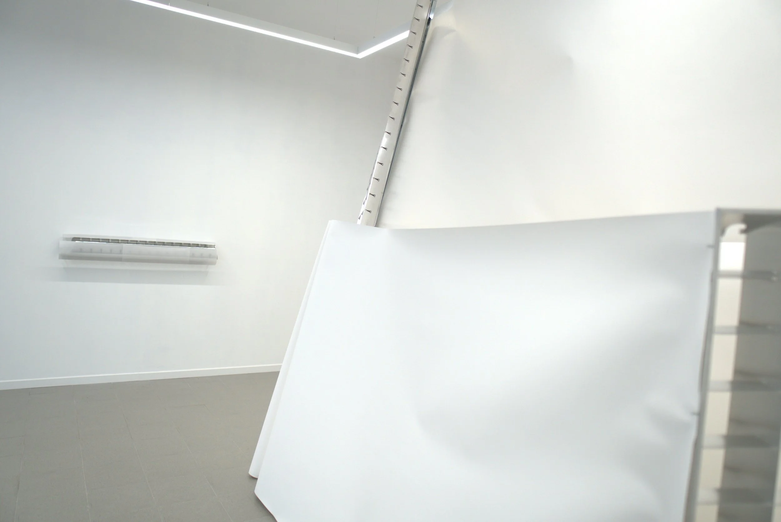 Interior de una sala con paredes blancas y una mesa blanca en primer plano. En la pared se observa un radiador y una estructura metálica inclinada en la esquina.