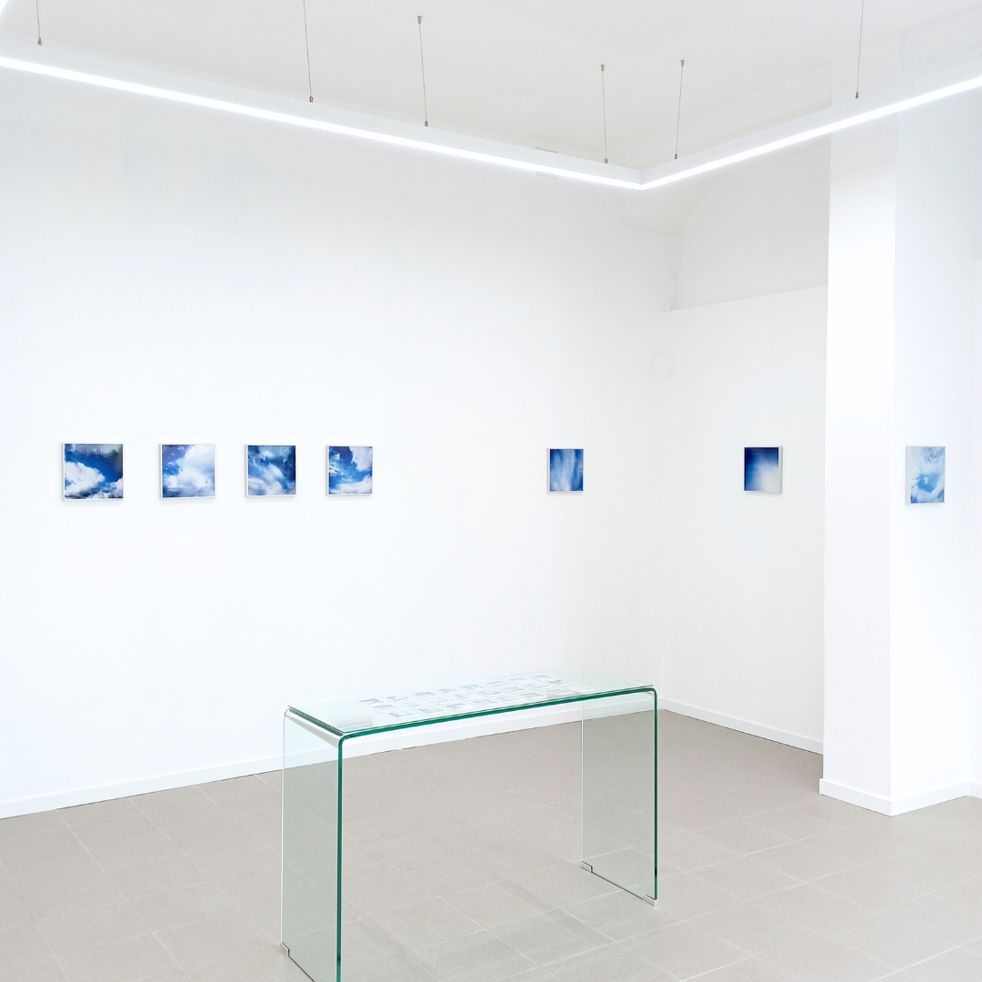 Galería con varias fotos de cielos con nubes en las paredes y un mueble de cristal en el centro.
