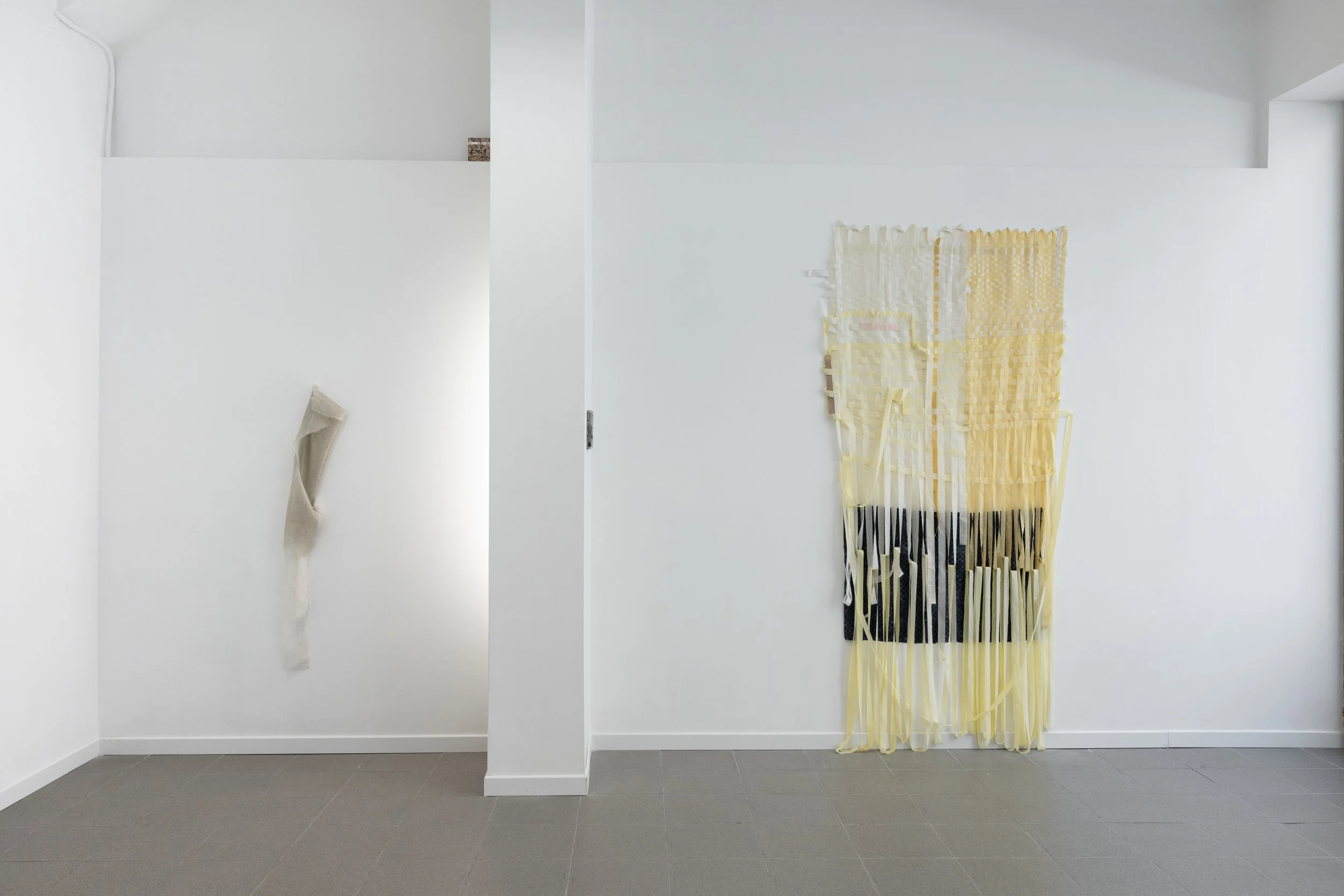 Instalación de arte con telas en tonos blancos y amarillos colgadas en una pared blanca en una galería moderna.