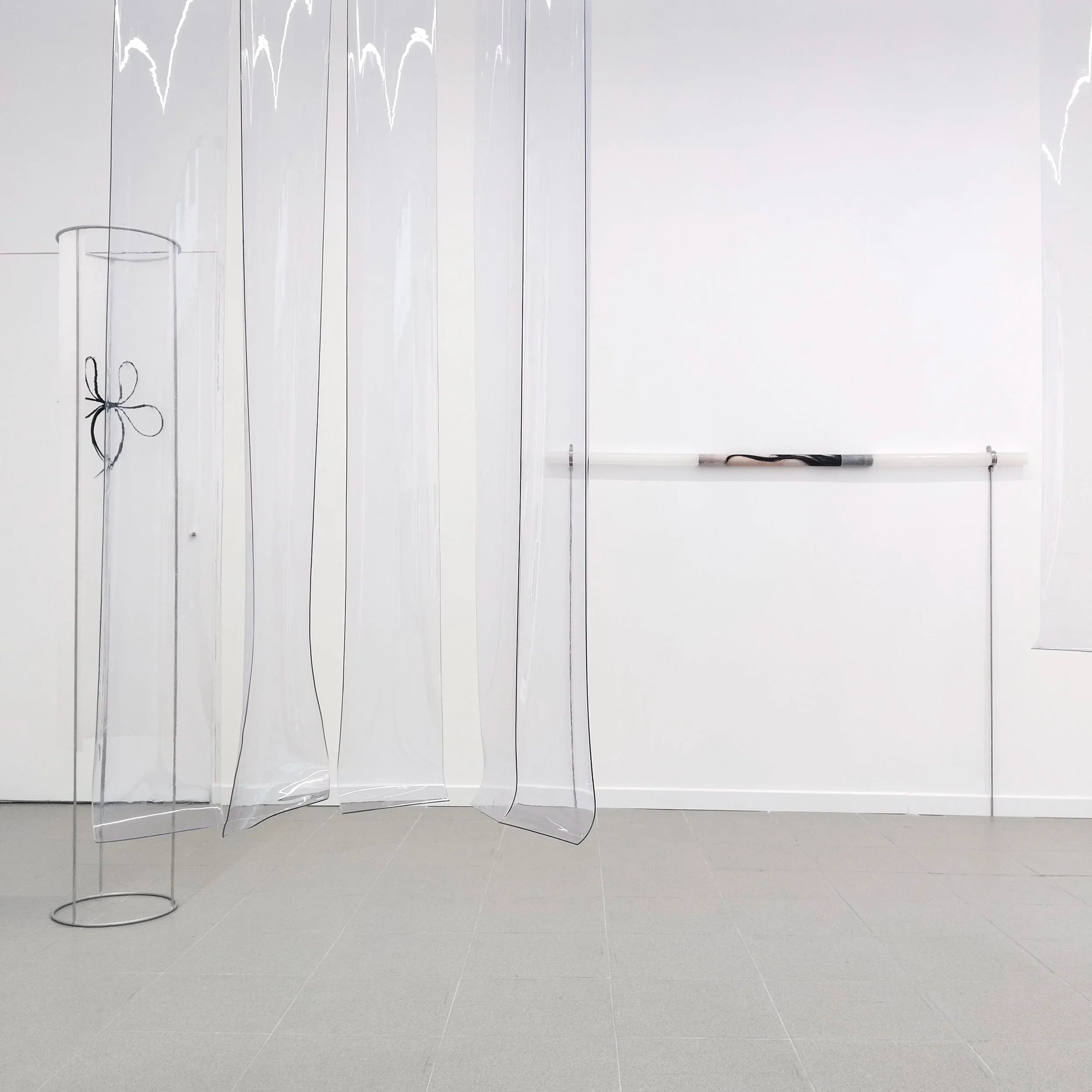 Espacio vacío con un fondo de paredes blancas y suelo gris. Se observan varias estructuras de acrílico transparente sujetas por soportes metálicos, con una de ellas amarrada con cinta negra en forma de lazo.