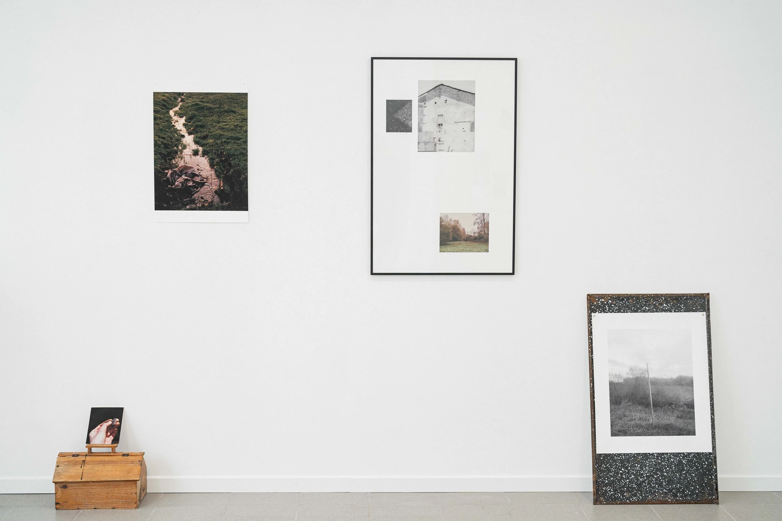 Muestra de varias obras de arte en una galería, incluyendo fotografías y cuadros en diferentes tamaños y estilos, con una caja de madera y una pantalla con un fondo de suelo gris.