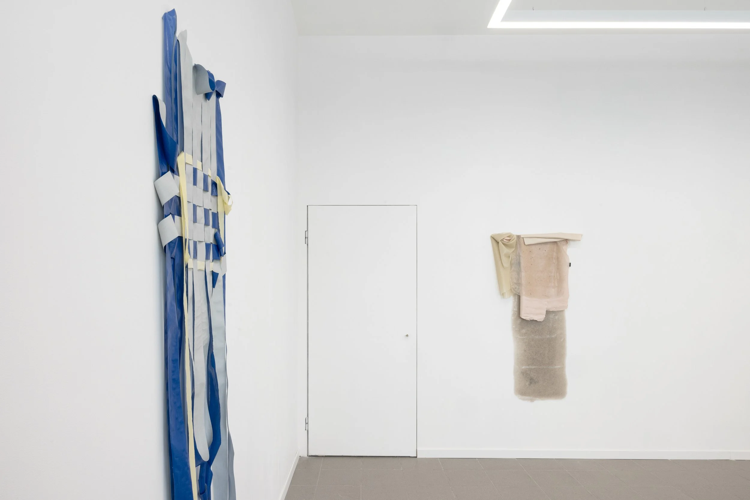 Instalación de arte contemporáneo con piezas textiles en tonos azul, gris y beige en una pared blanca de galería