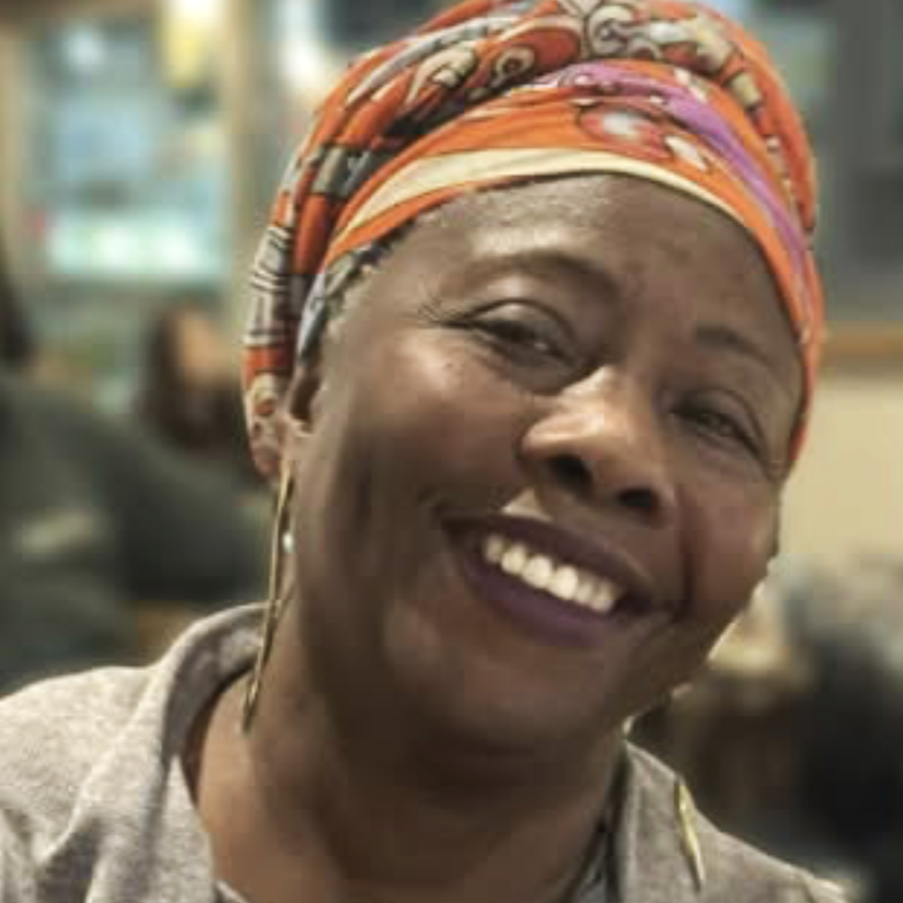 A smiling woman wearing a colorful headscarf and hoop earrings, in a lively indoor setting.