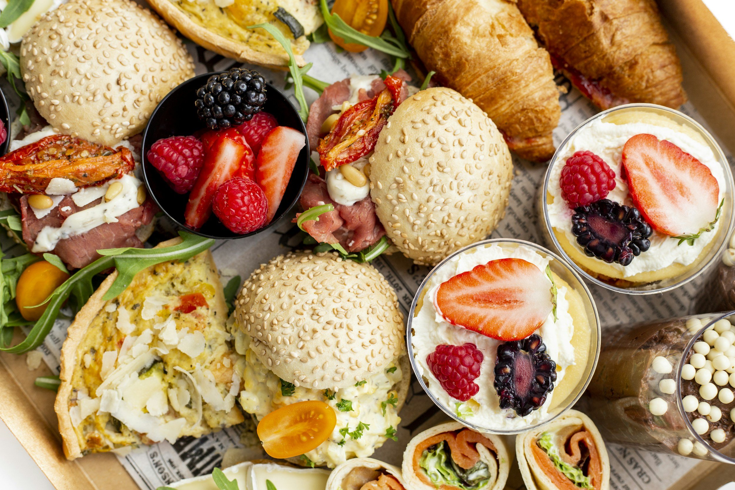 An assortment of finger foods including mini sandwiches, wraps, fruit cups, and croissants on a serving tray.