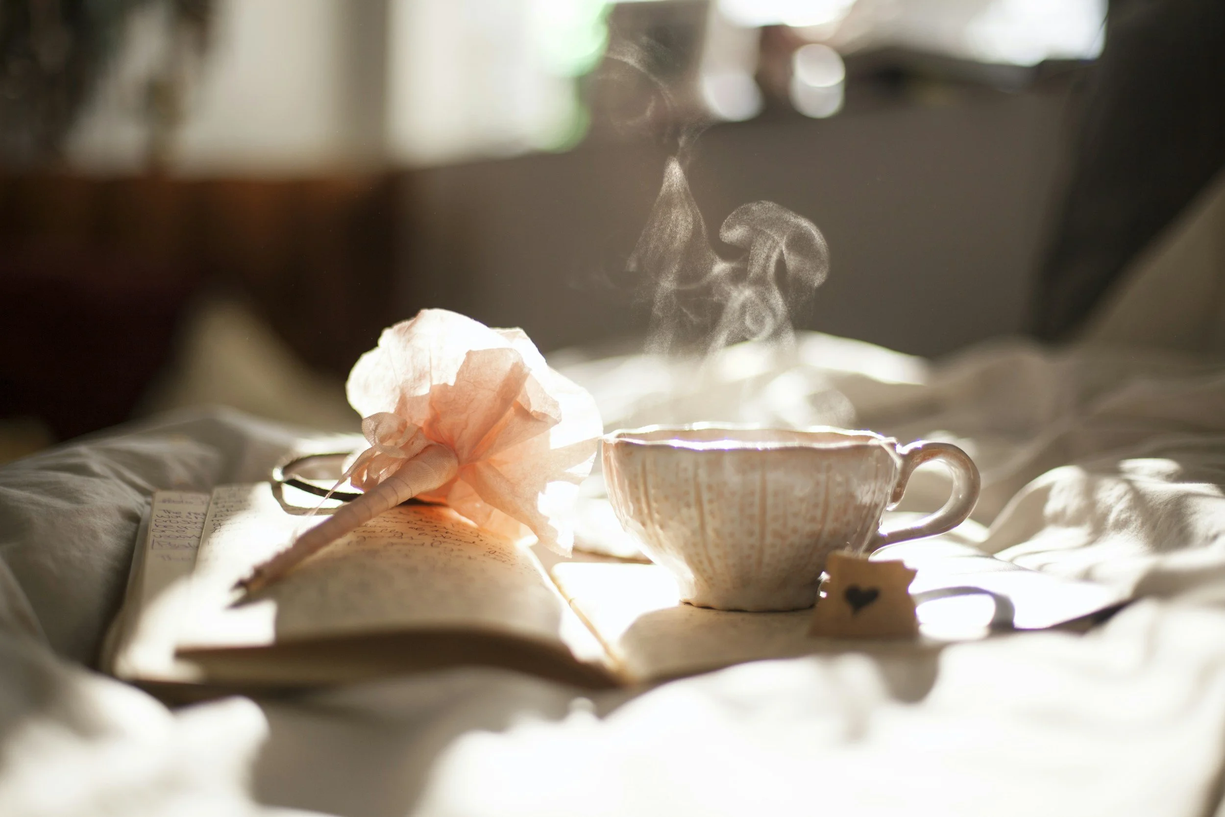 A steaming cup of tea on a white cloth with soft natural light, a pink tissue, and an open notebook with handwritten notes nearby.