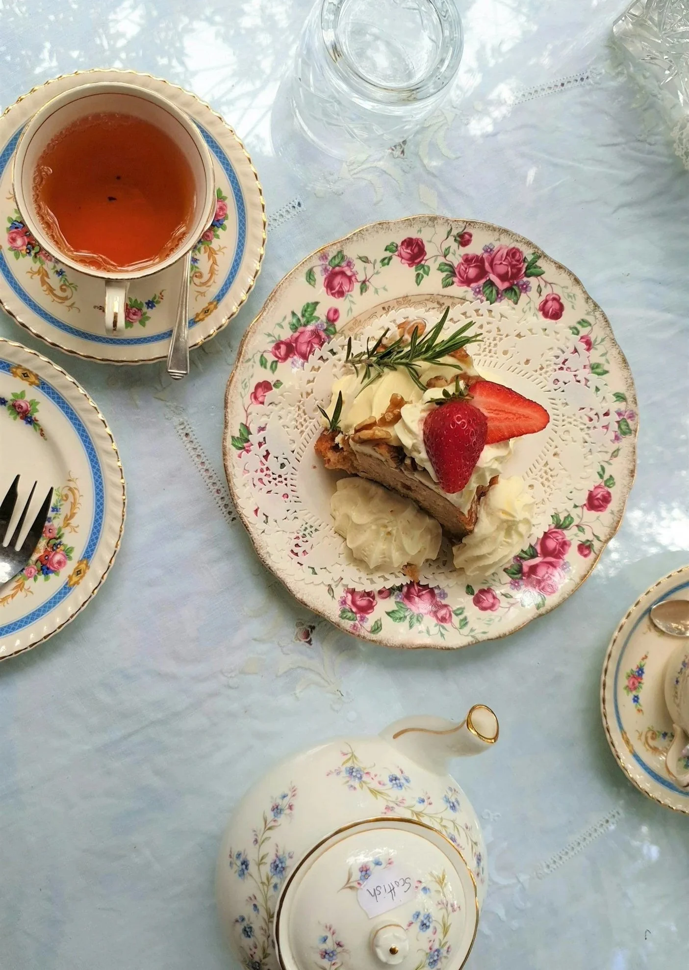 A table set with ornate floral china featuring a cup of tea, a glass of water, and a slice of cake topped with strawberries and garnished with herbs, on a light-colored tablecloth.