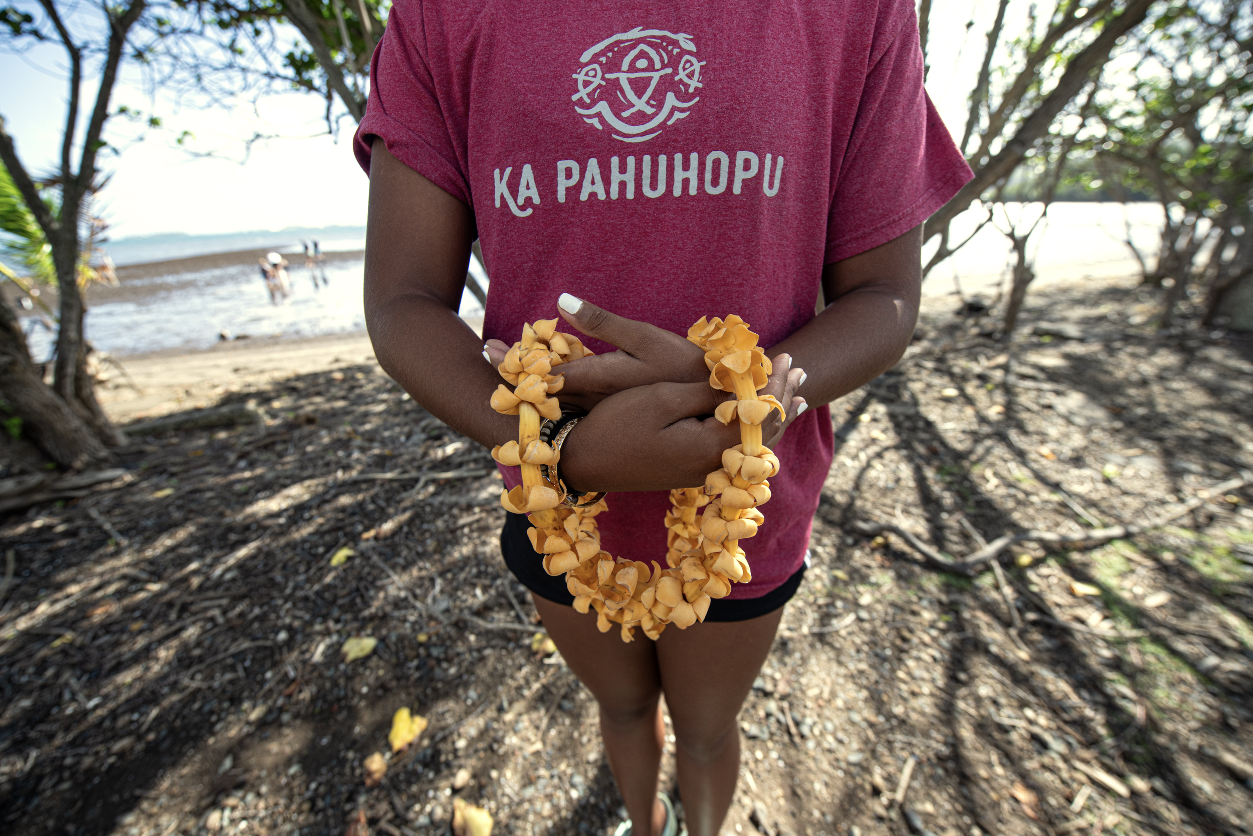 Person holding a yellow flower lei at the beach, wearing a maroon shirt with white text and a graphic, black shorts, and bracelets, with trees and beach in the background.
