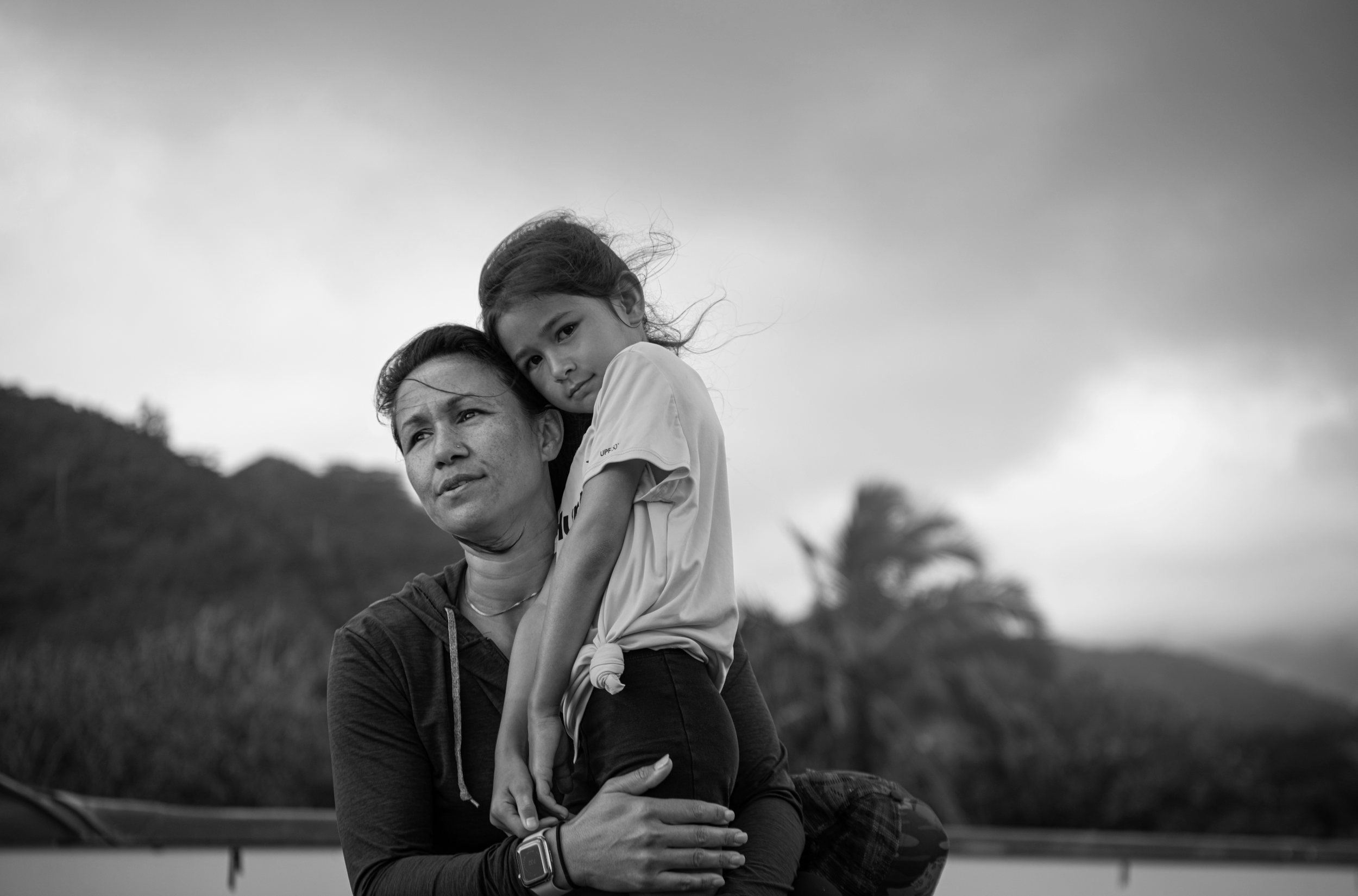 A woman carrying a young girl on her back outdoors, with trees and hills in the background, black and white photo.