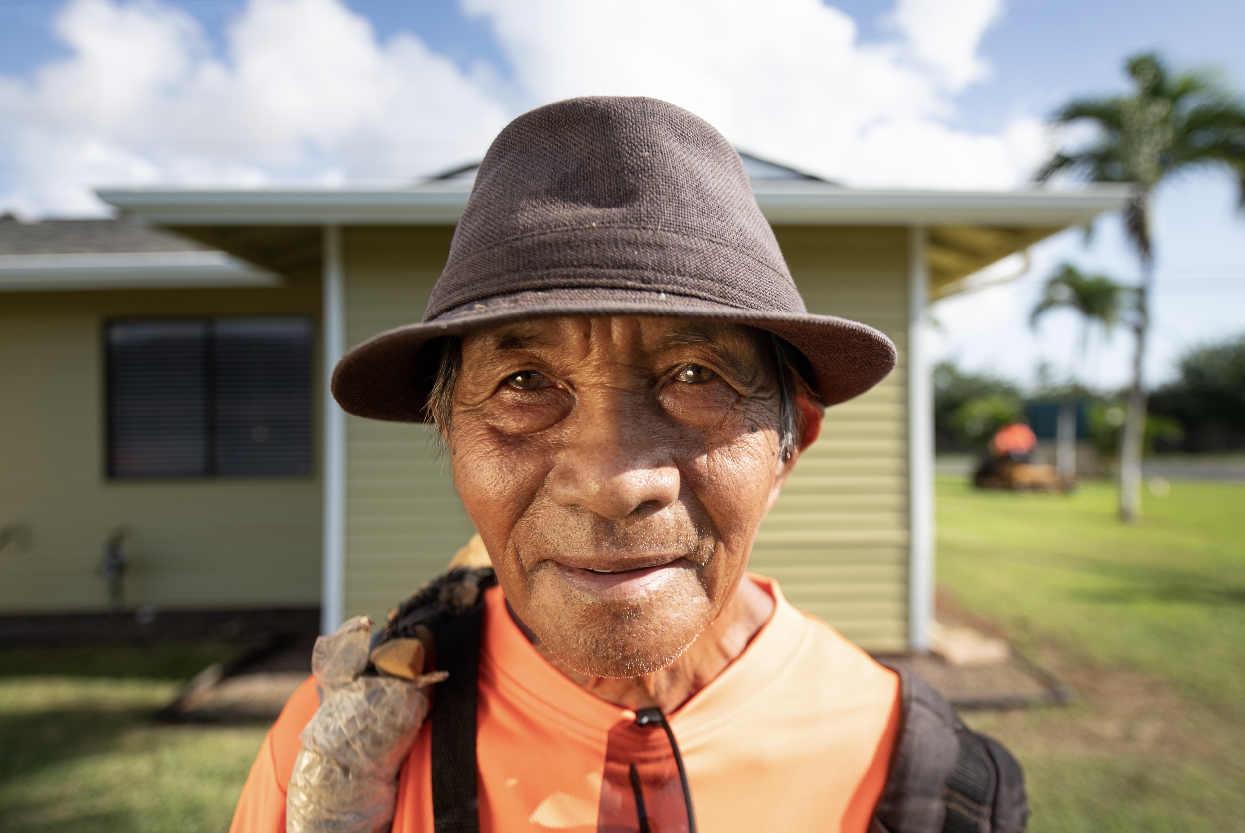 Close-up of an elderly man wearing a brown hat and orange shirt, standing outdoors in front of a yellow house with trees and a partly cloudy sky in the background.