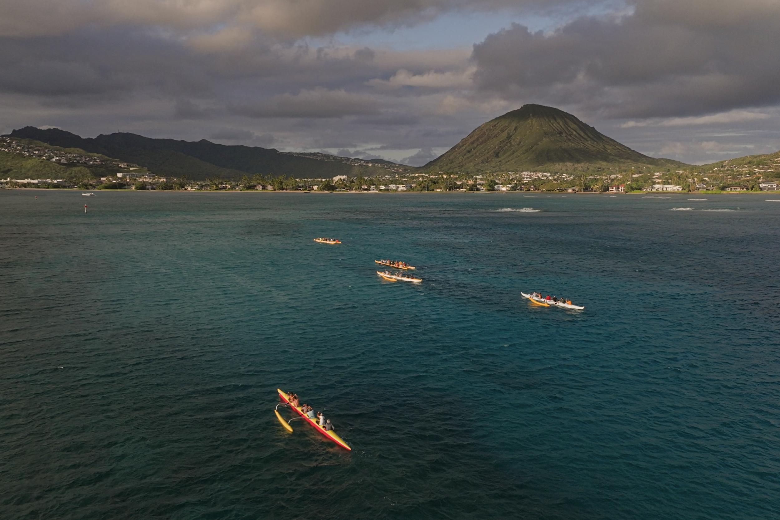 Several boats on blue ocean water near shoreline with a mountain in the background, cloudy sky overhead.