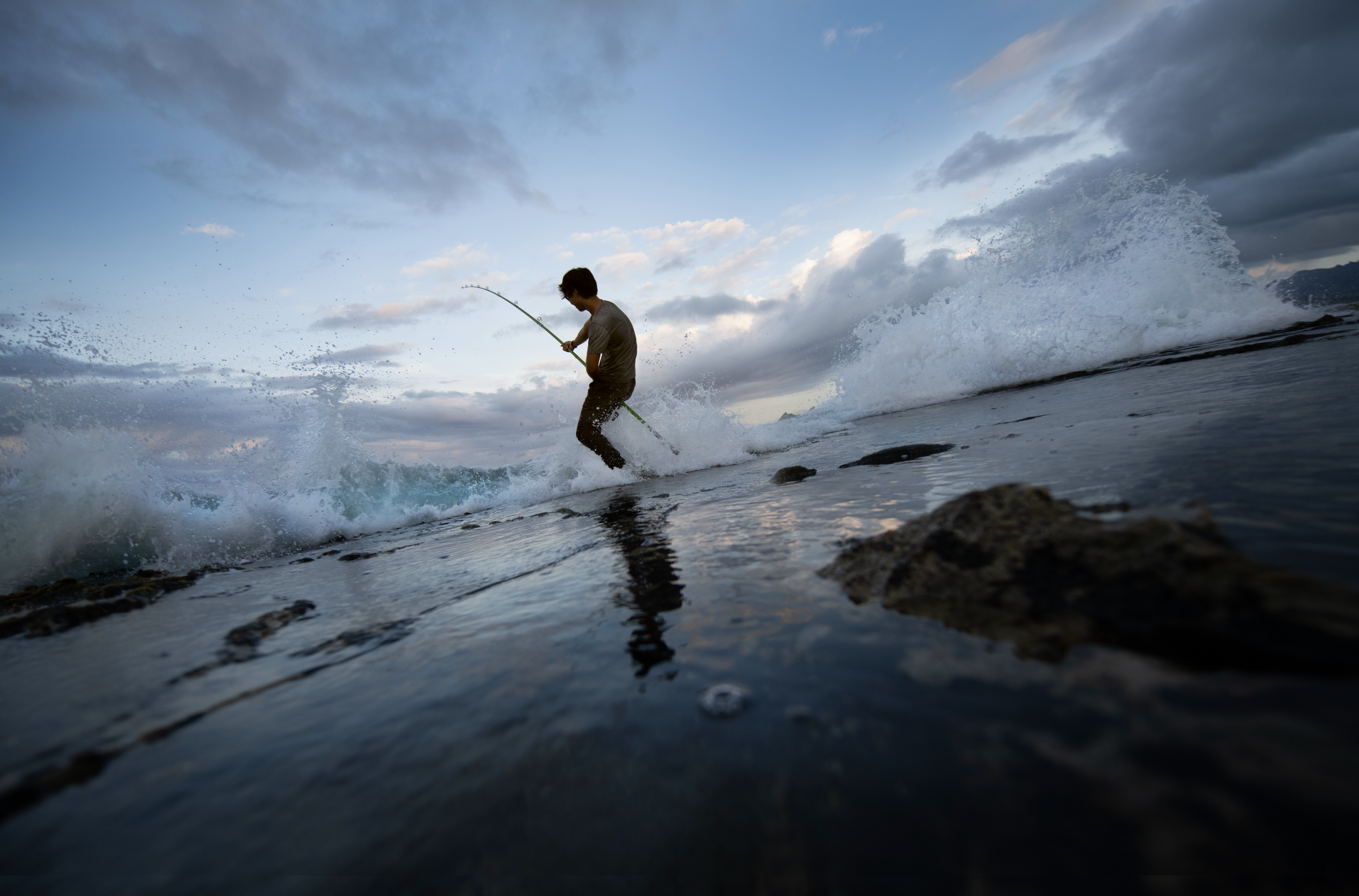 Person fishing on a rocky shoreline during sunset with cloudy sky and waves splashing.