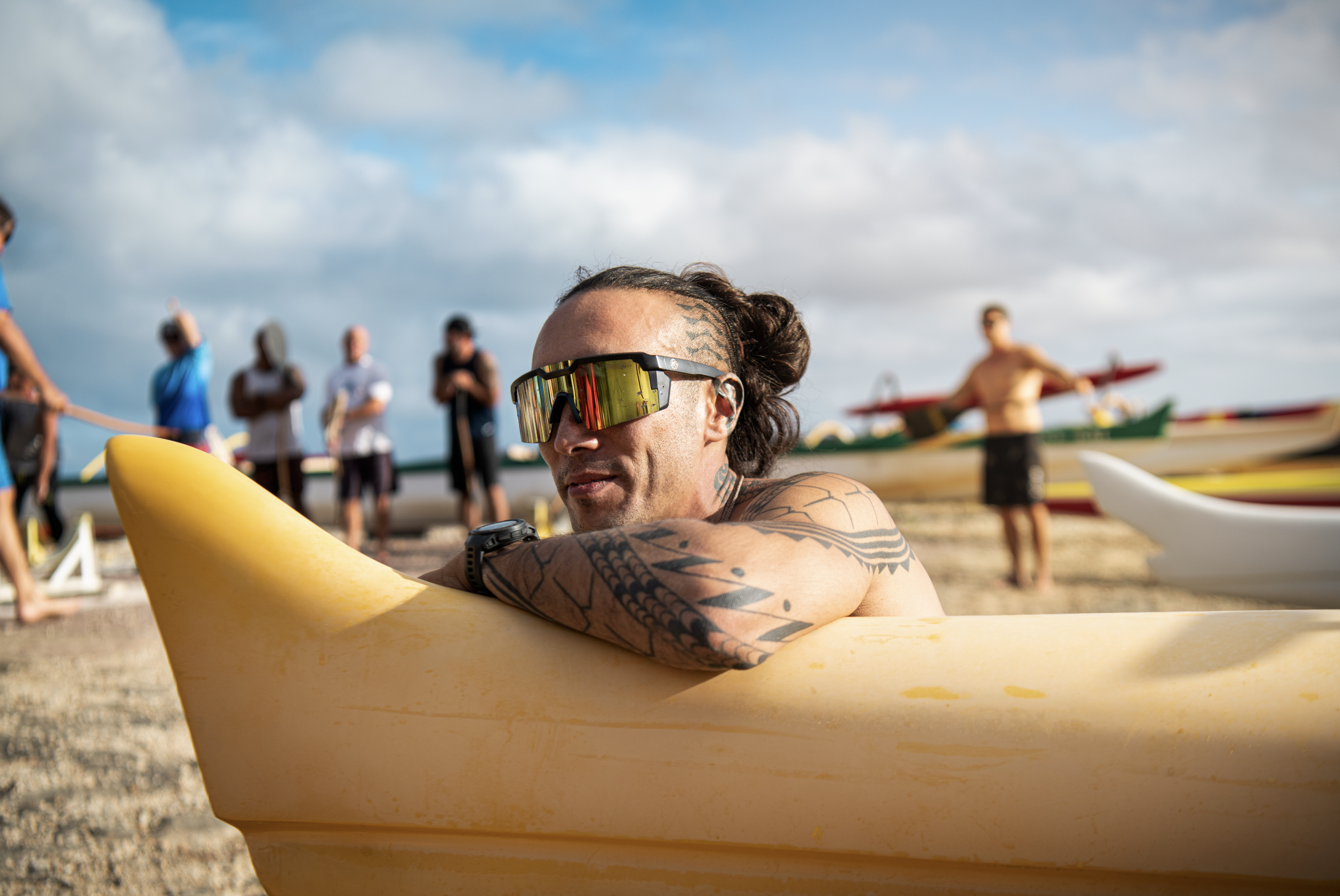 Tattooed man with sunglasses resting on yellow kayak at the beach, with people and kayaks in the background under cloudy sky.