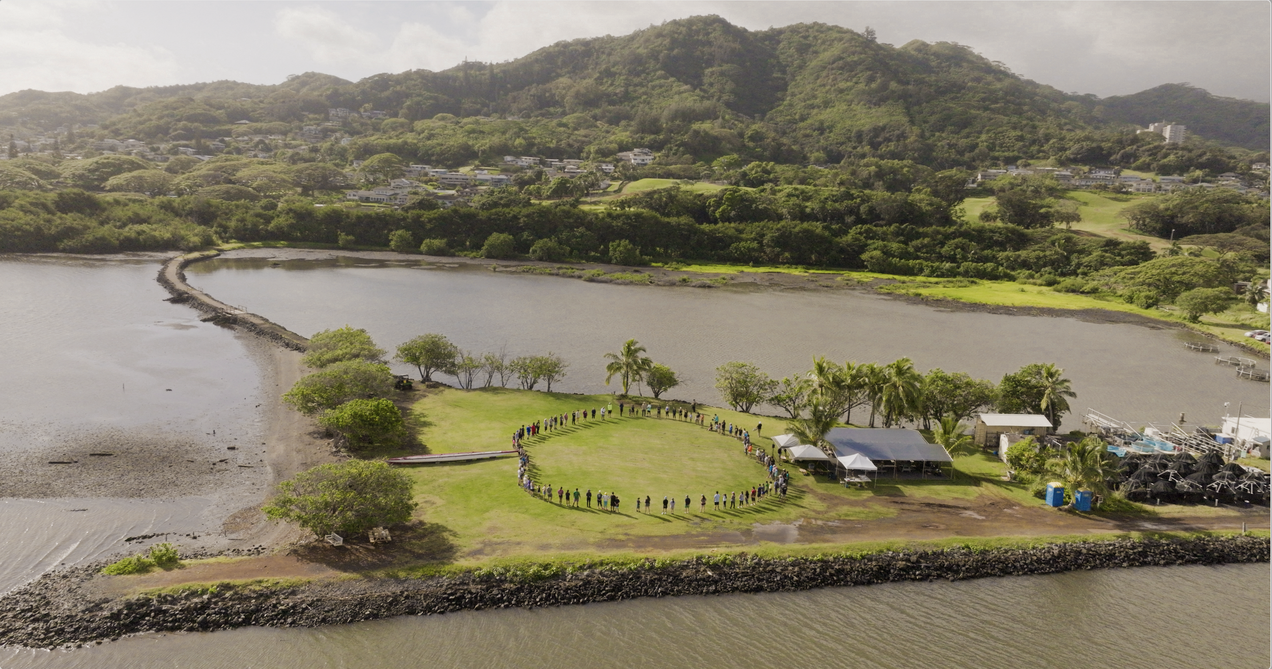 Group of people standing in a circle on a grassy area near a waterfront, surrounded by trees and a few small buildings, with hills and residential areas in the background.