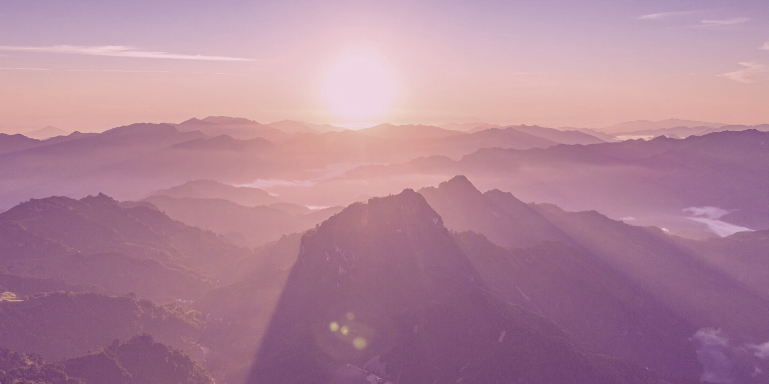 Sunrise over misty mountain peaks with pink and purple hues, casting shadows and light beams through the clouds.