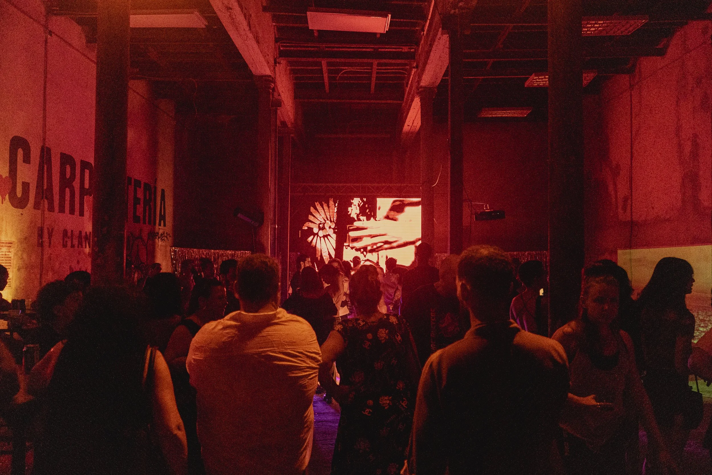 Un gran grupo de personas sentadas en una sala oscura con una pantalla grande al fondo, en un entorno que parece ser un evento o concierto con iluminación roja.