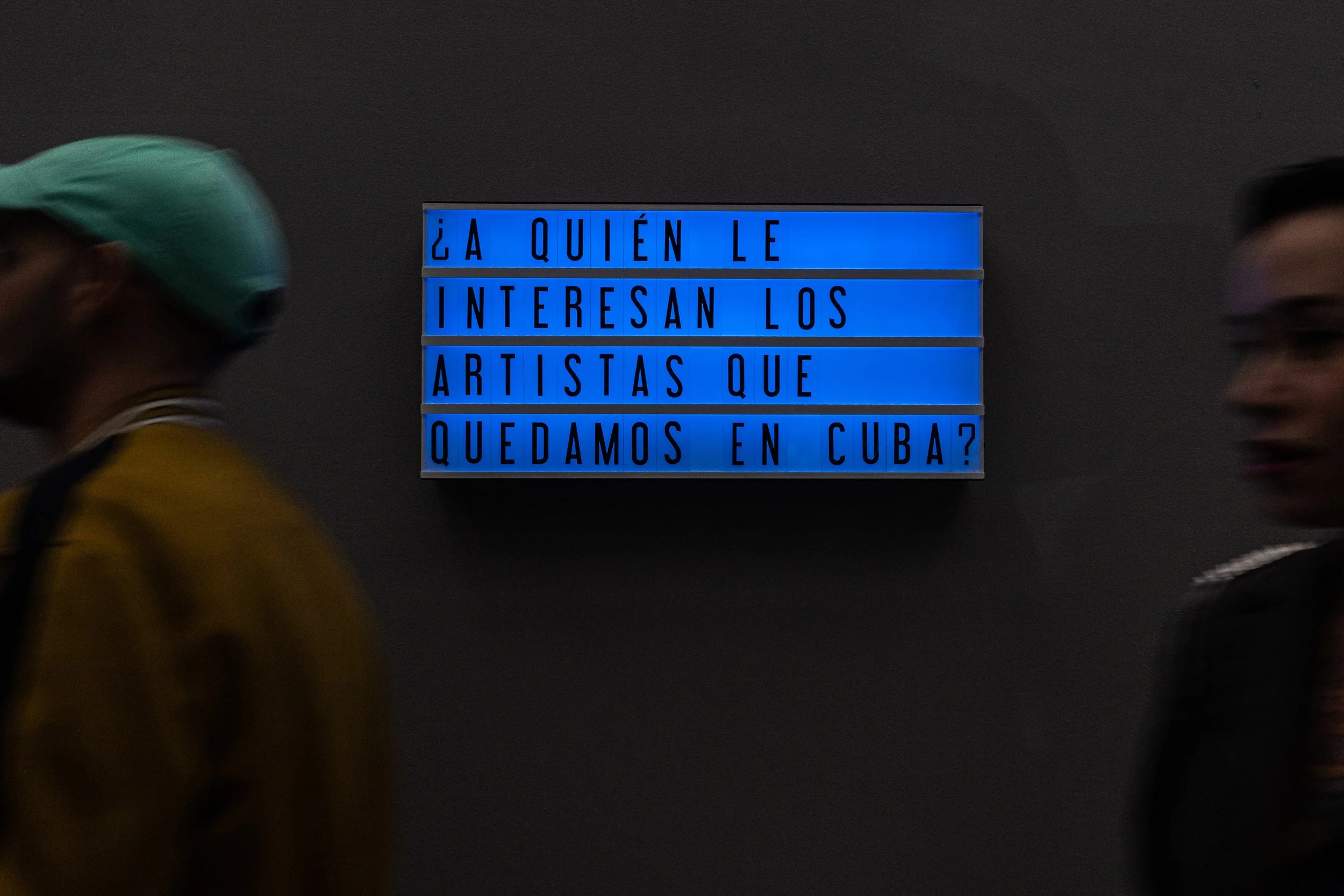 Una señal de luces azules con letras negras que dice: "¿A quién le interesan los artistas que quedamos en Cuba?" en un entorno oscuro, con personas borrosas que pasan frente a ella.
