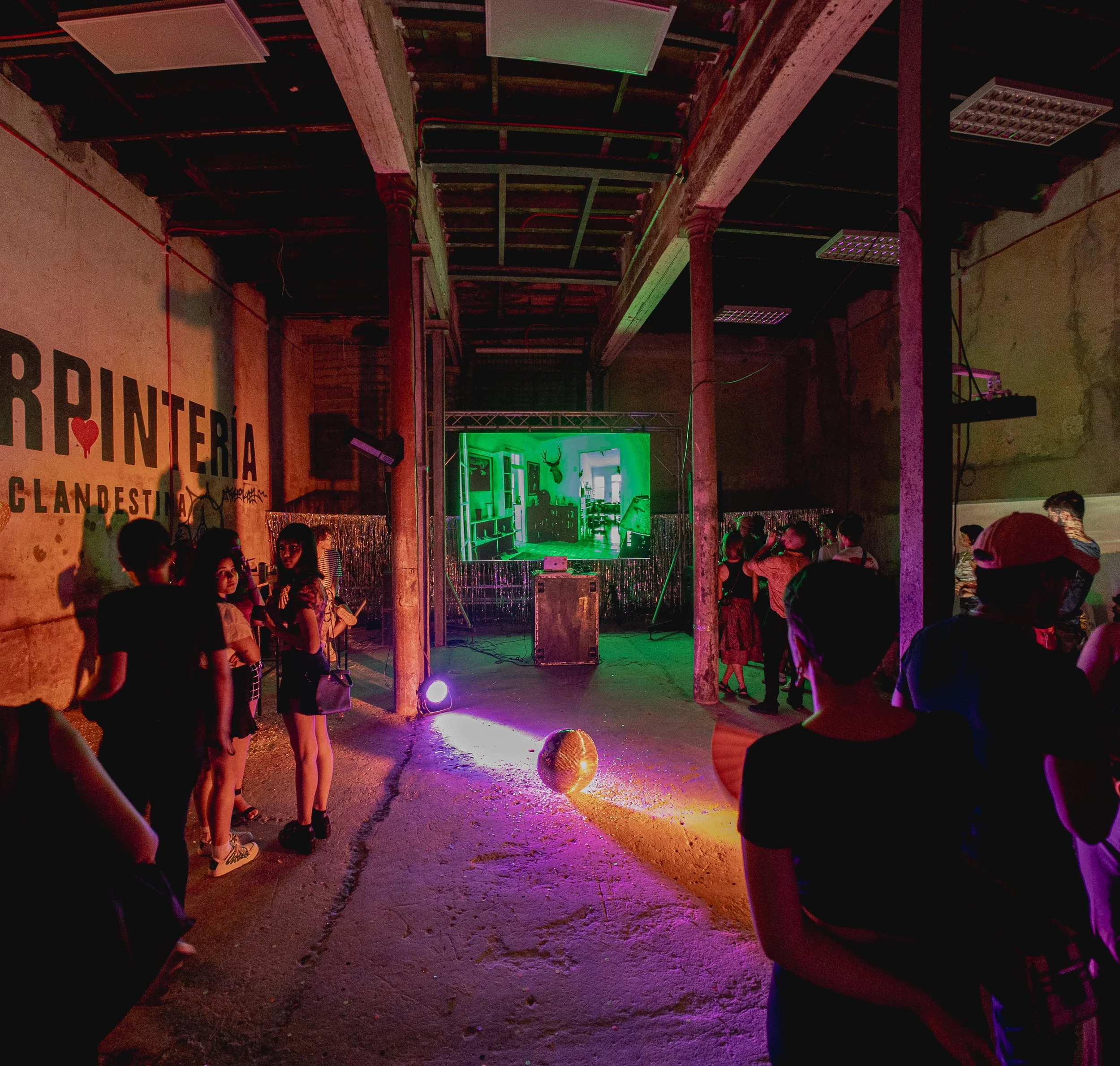 Un grupo de personas en un espacio abierto con iluminación teatral, paredes en proceso de construcción y una pantalla grande al fondo, en un ambiente que parece un evento o presentación cultural.