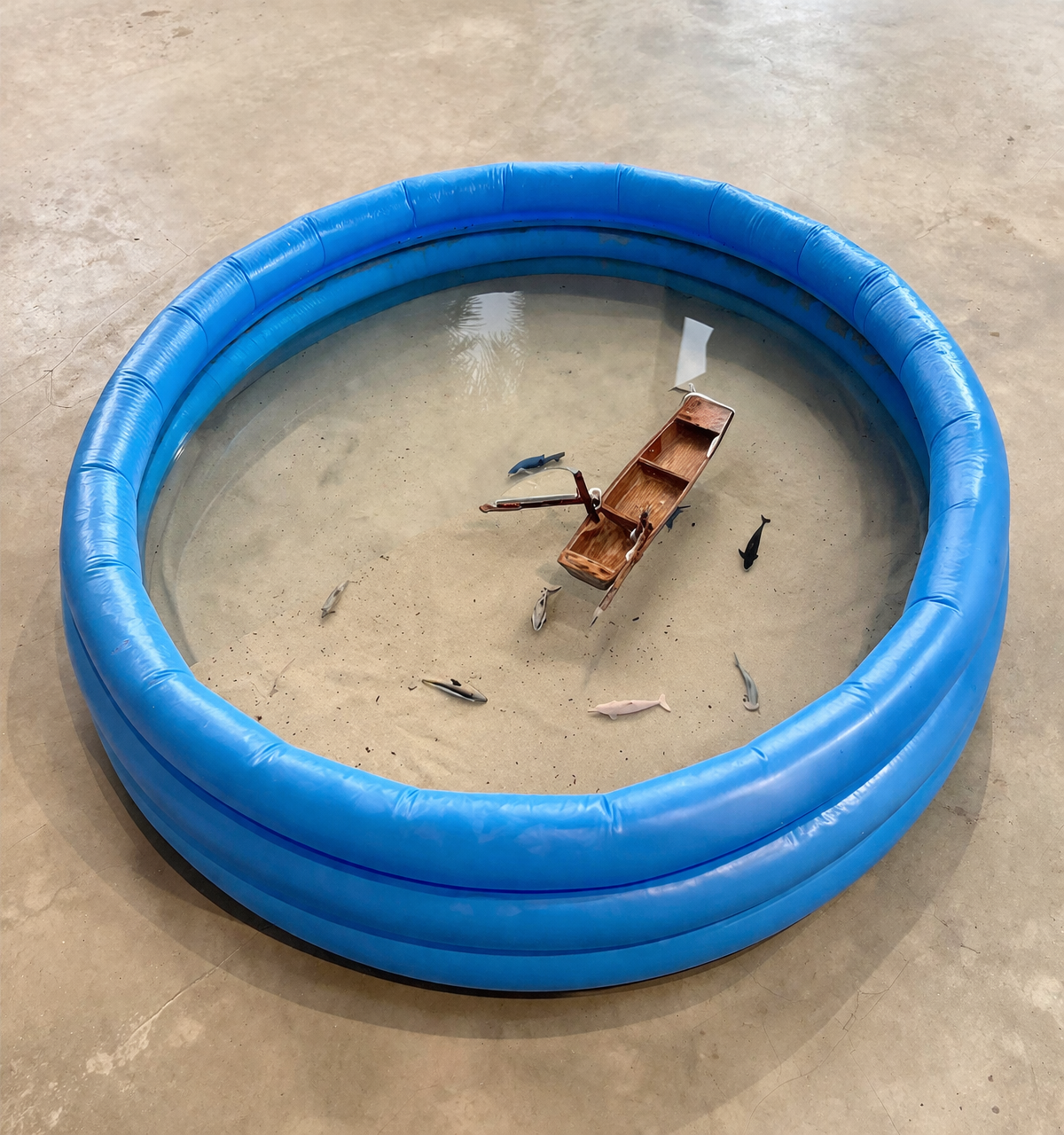 Una piscina inflable azul con agua y peces de plástico flotando en ella, junto a una pequeña embarcación y una caña de pescar.