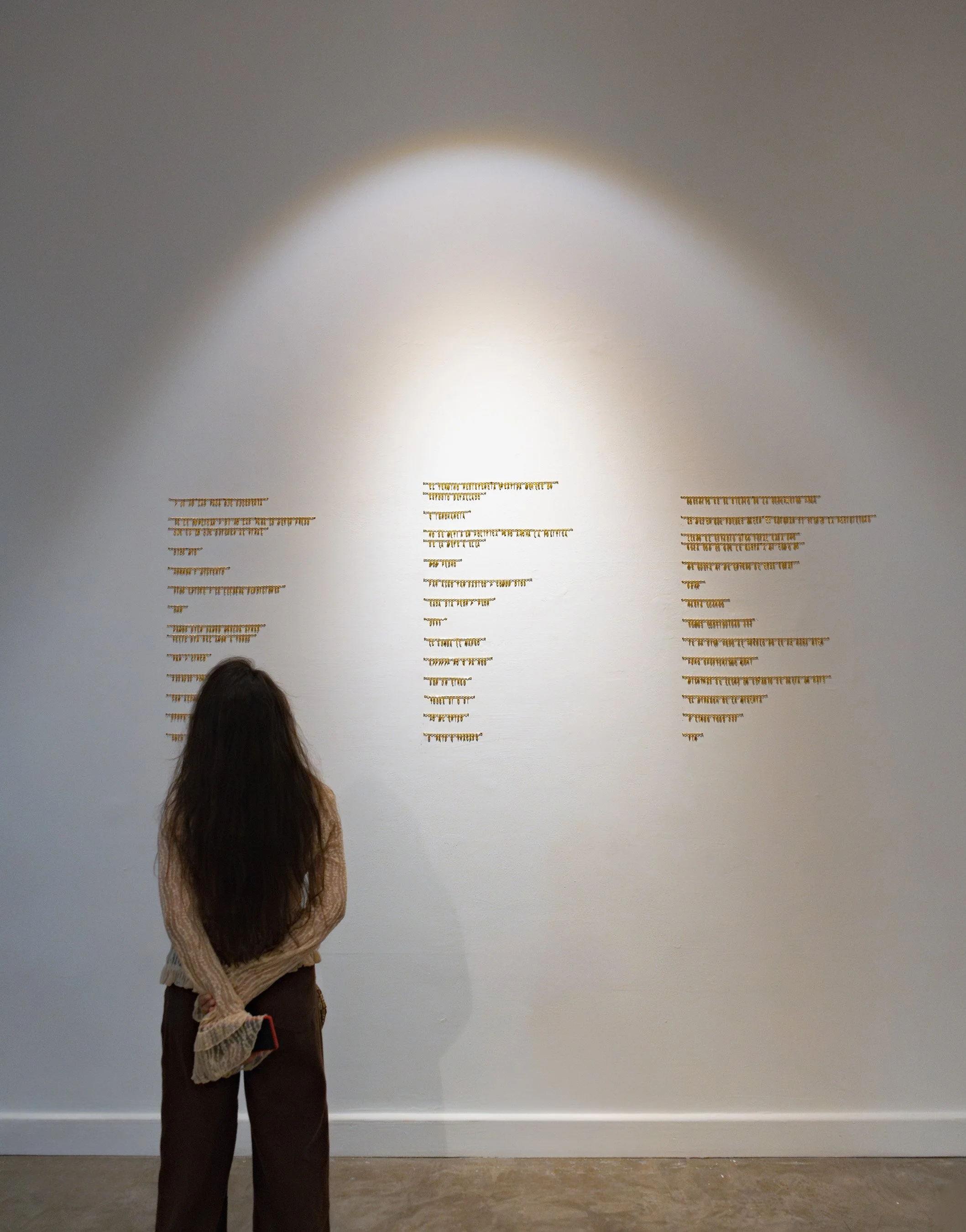Una mujer con cabello largo, camiseta beige y pantalones oscuros observa una pared blanca con textos en dorado. La luz dirigida ilumina la pared y la mujer.