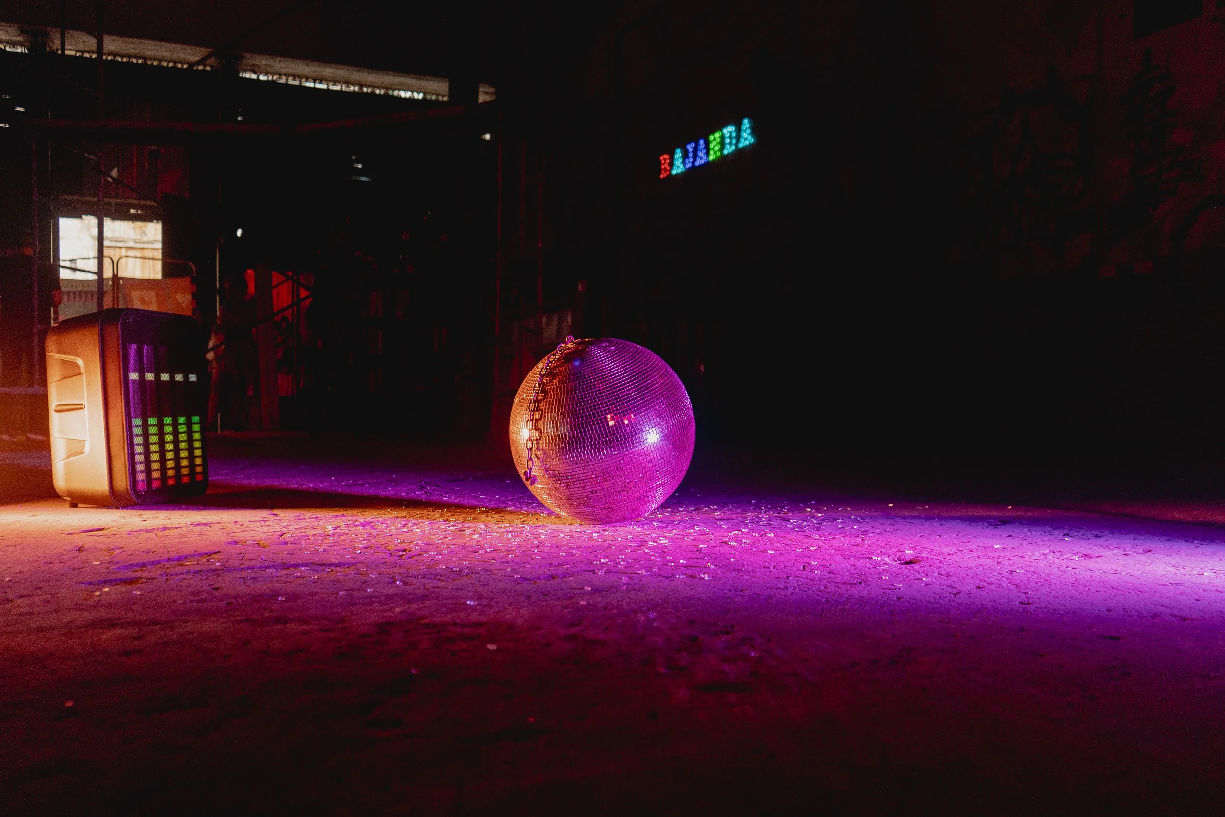 Una bola de discoteca brillante, una caja de sonidos con pantalla LED y el cartel de 'Bar' en un ambiente oscuro con luces de colores, típicos de una fiesta o club.