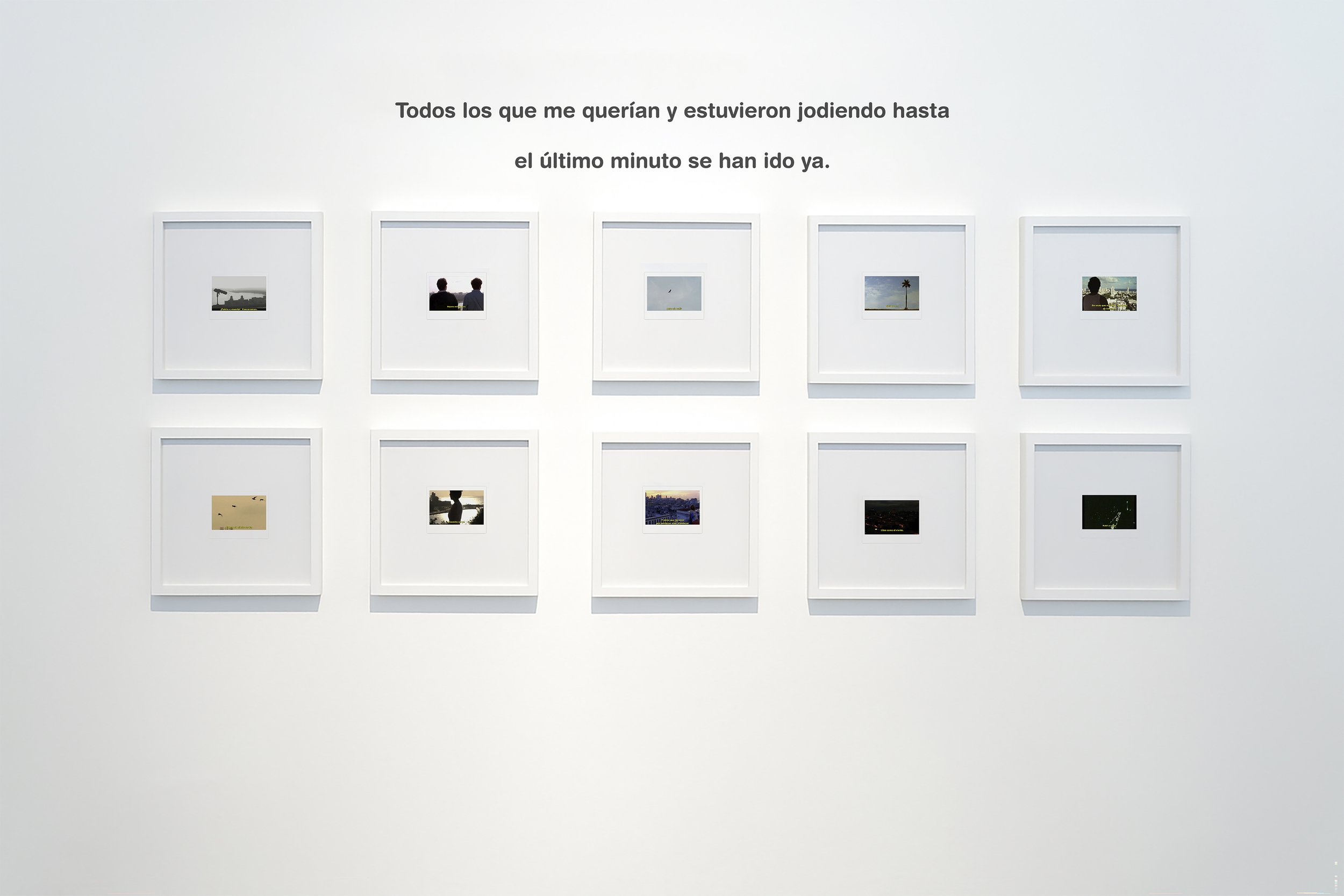 Exposición de varias pequeñas fotografías en marcos blancos en una pared blanca, con un texto en español en la parte superior quedice: 'Todos los que me querían y estuvieron jodiendo hasta el último minuto se han ido ya.'