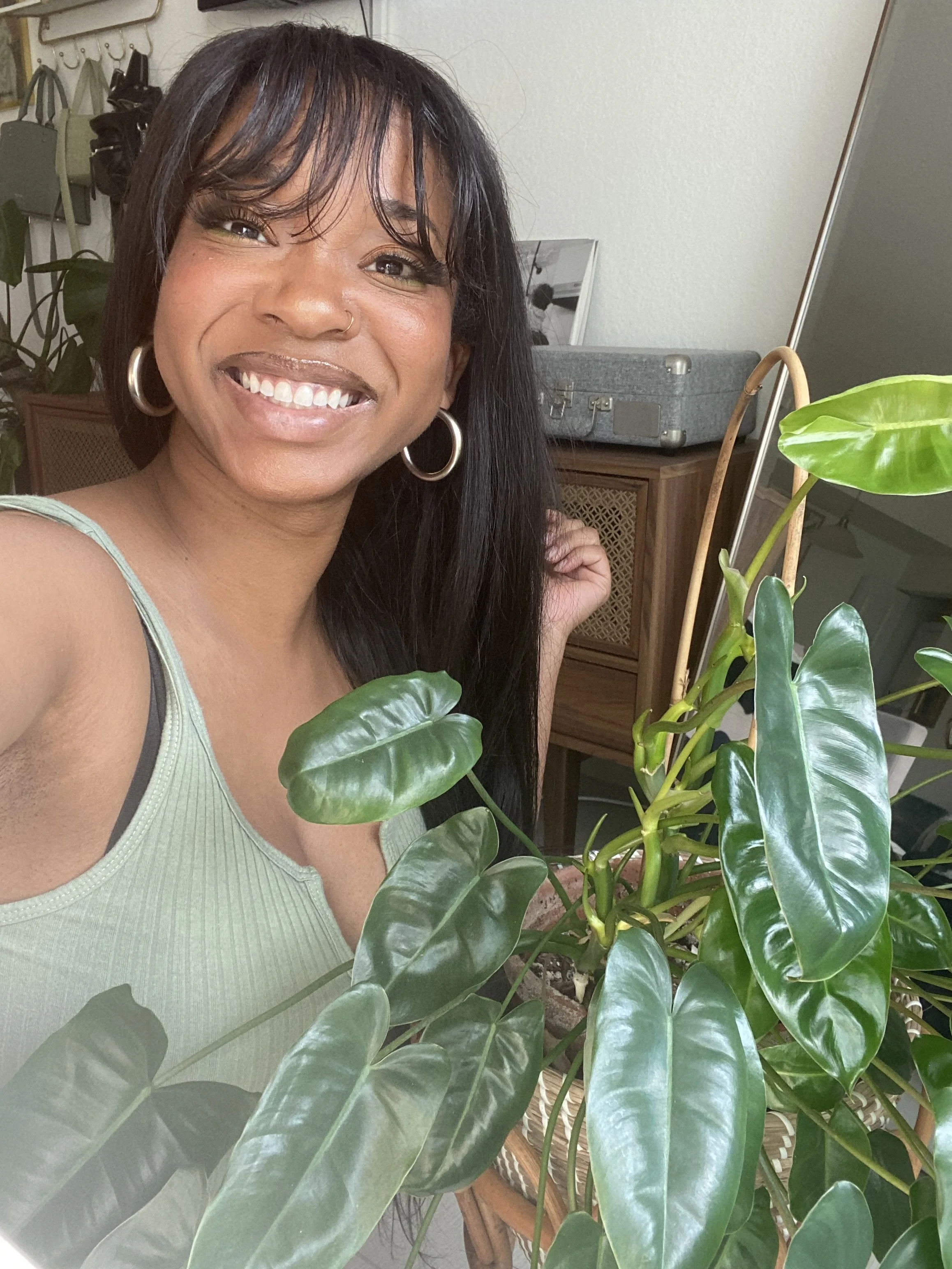 A woman with long black hair smiling, wearing a light green tank top and hoop earrings, standing behind a large green leafy houseplant in a room with a wooden cabinet, a gray suitcase, and a mirror.