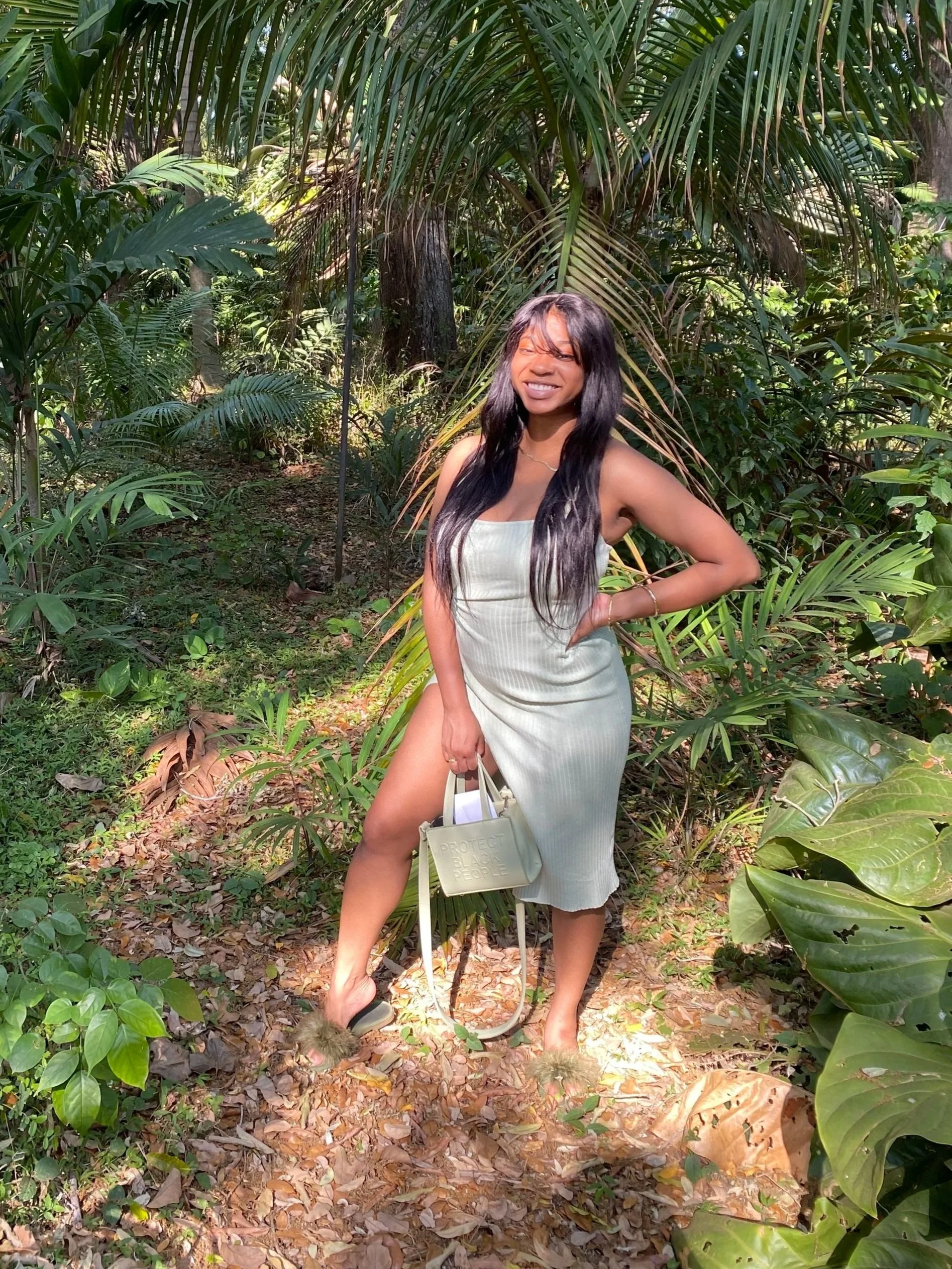 A woman in a light green dress with a slit stands on a leaf-covered path in a lush, green jungle, smiling at the camera while holding a beige handbag.