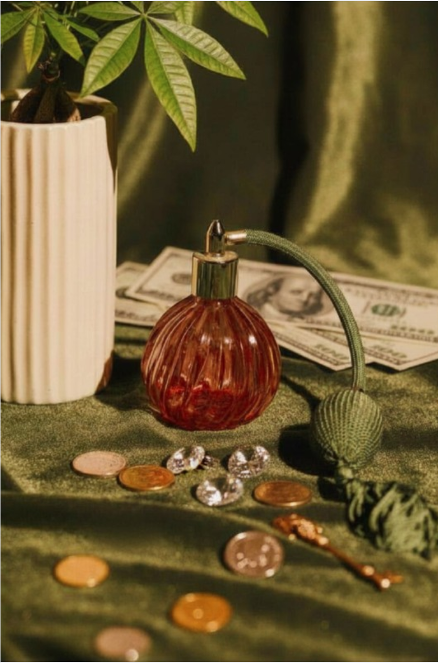 A vintage-style perfume bottle with a green tassel and button, a potted plant, U.S. dollar bills, and scattered coins and jewelry on a green fabric background.