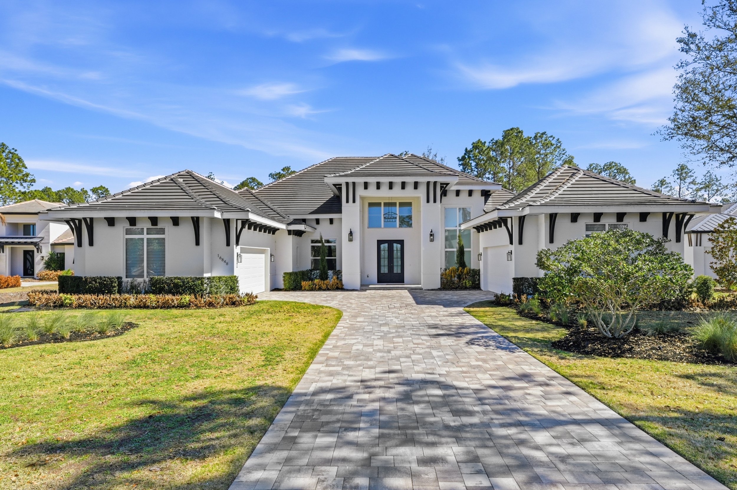 Large modern white house with black accents and a gray tiled roof, front yard with greenery and landscaping, paved walkway leading to the front door, blue sky with some clouds.
