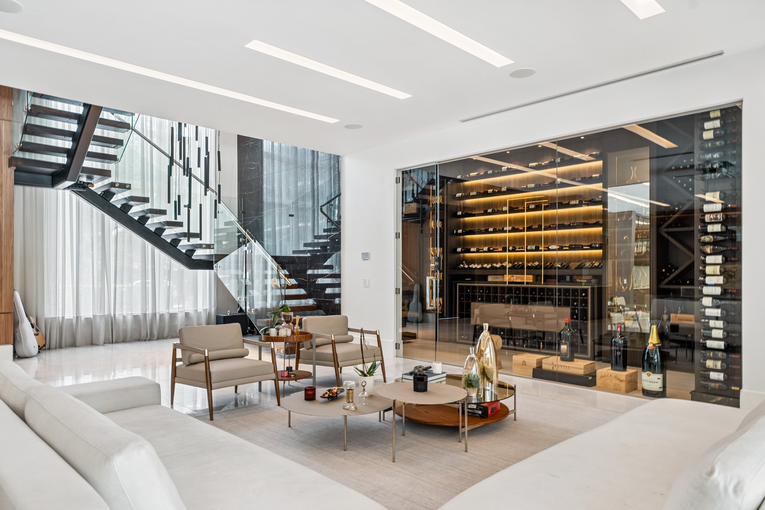 Modern living room with glass staircase, a large wine cellar with bottles displayed on illuminated racks, beige sofas, and contemporary decor.