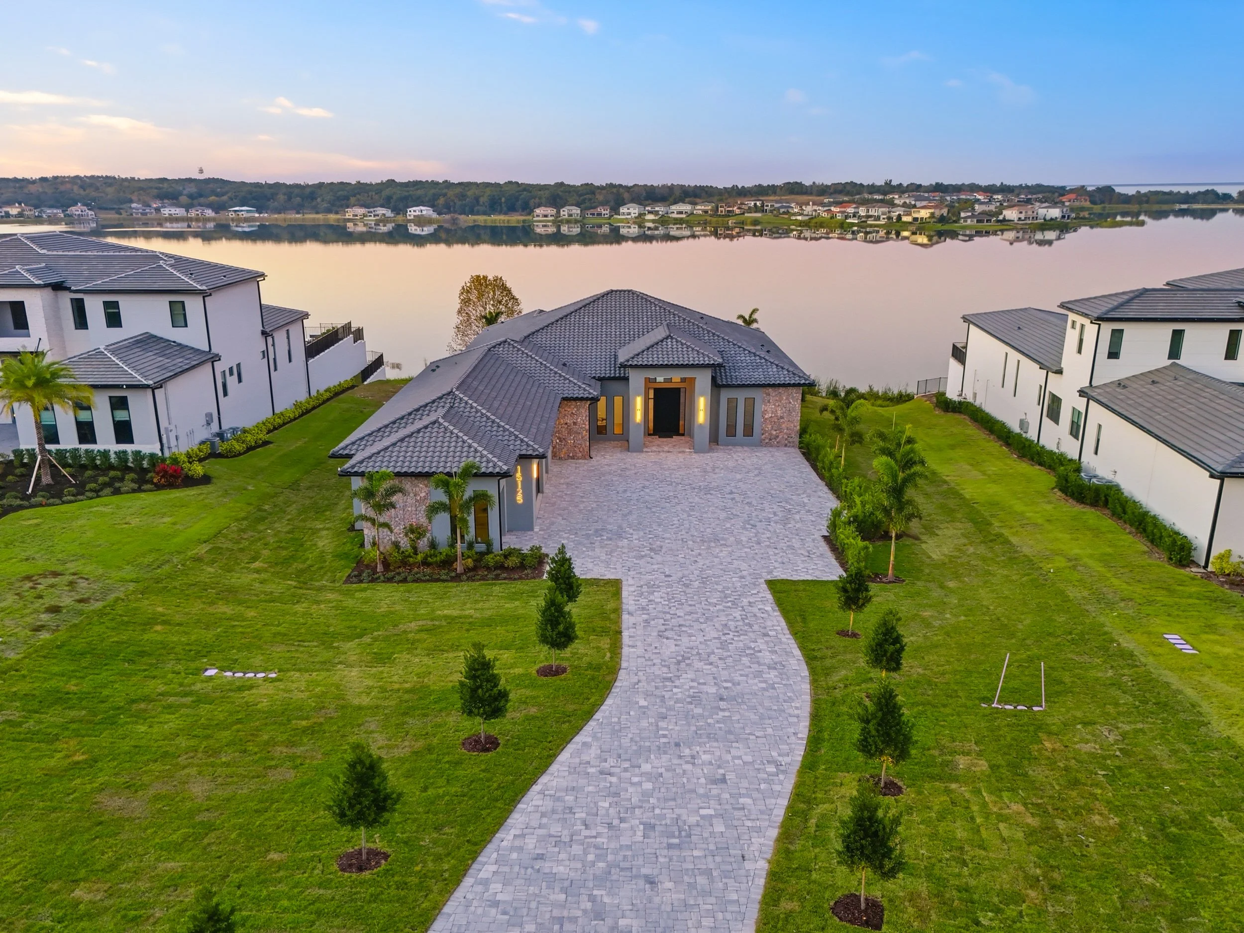 A modern house with a paved driveway, green lawn, and small trees, situated near a calm lake.