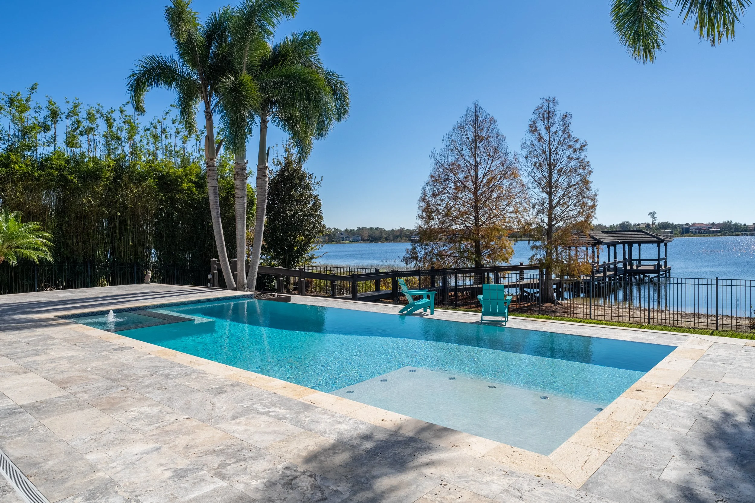 Swimming pool with a water feature, overlooking a lake, with trees and a wooden dock in the background, under a clear blue sky.