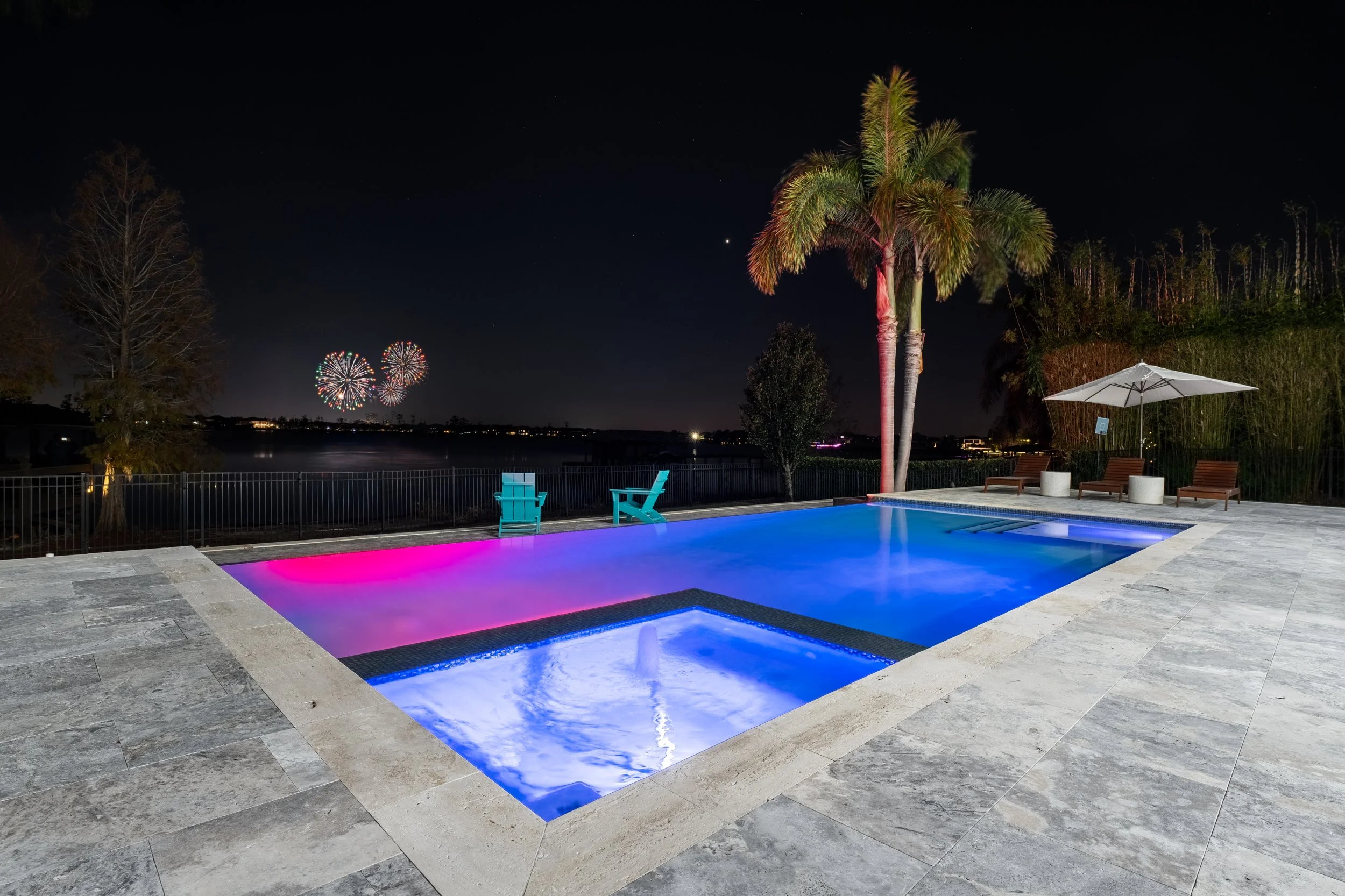 Nighttime view of an illuminated swimming pool with colorful LED lights, surrounded by patio furniture, palm trees, and fireworks in the sky.