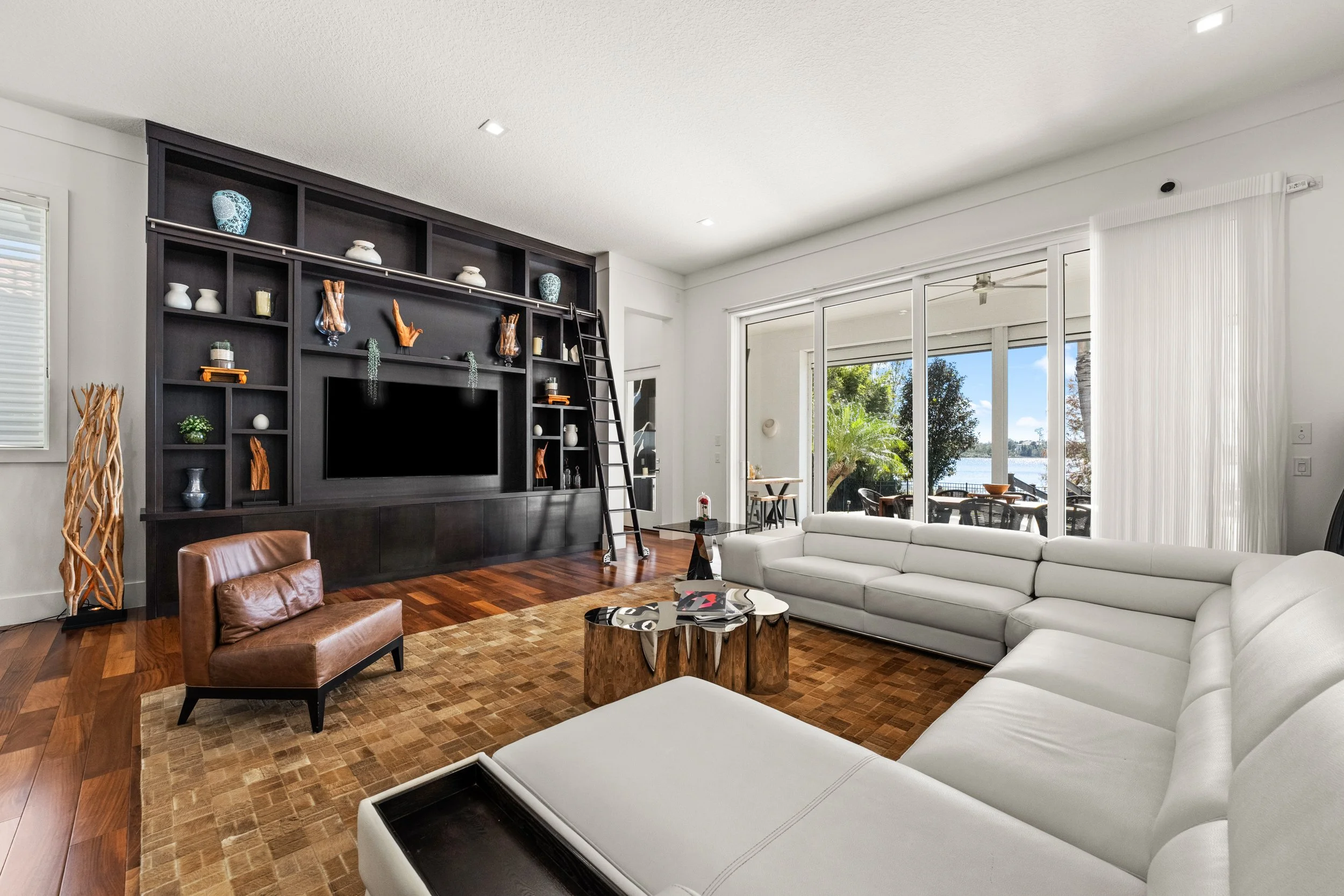 Living room with a large white sectional sofa, a brown leather chair, a round nesting table, a black entertainment center with shelves and a ladder, and glass sliding doors leading to an outdoor patio with trees and water view.