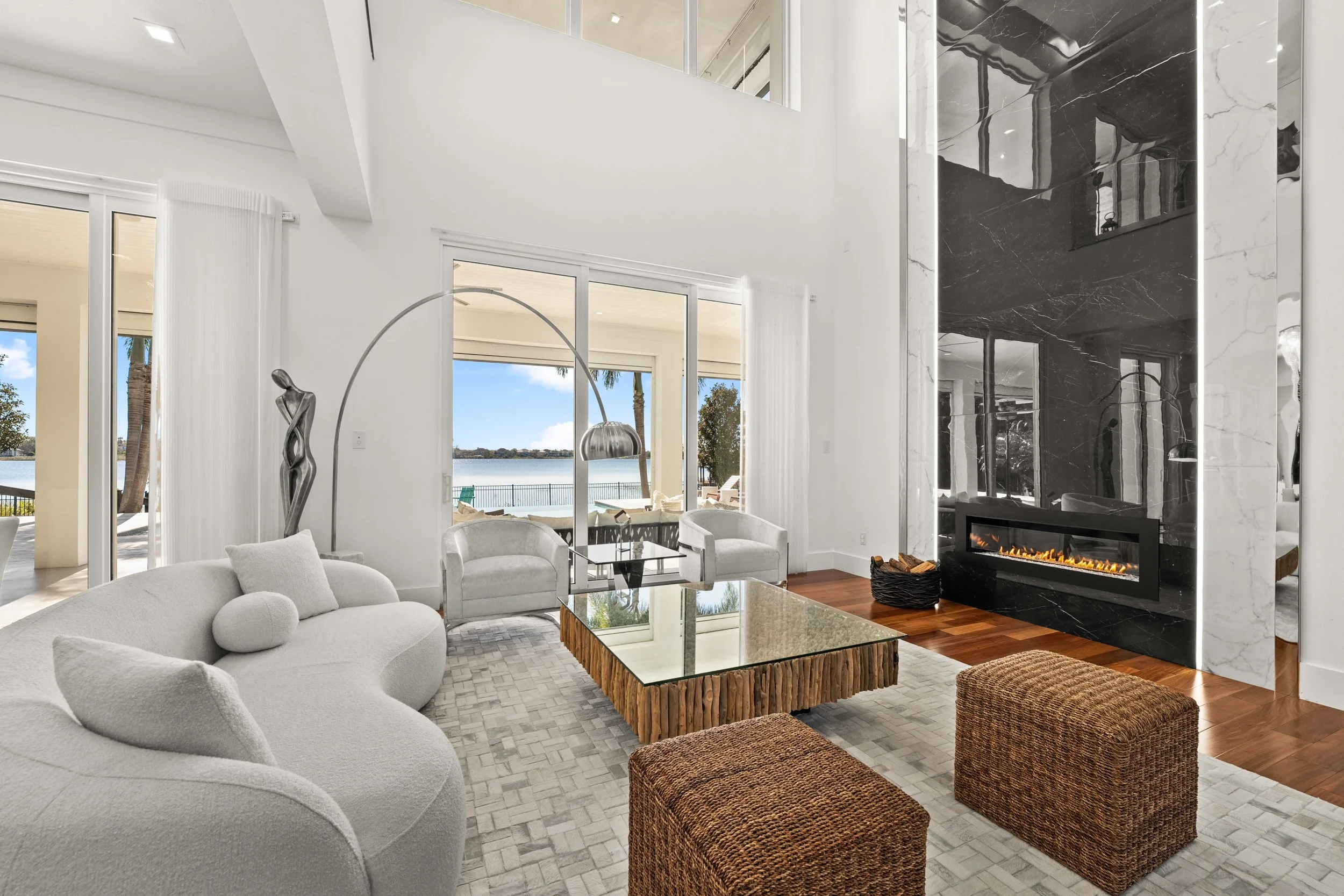 Modern living room with a white curved sofa, glass-top coffee table with a wooden base, two woven ottomans, and a black marble fireplace, large sliding glass doors showing a patio and view of water outside.